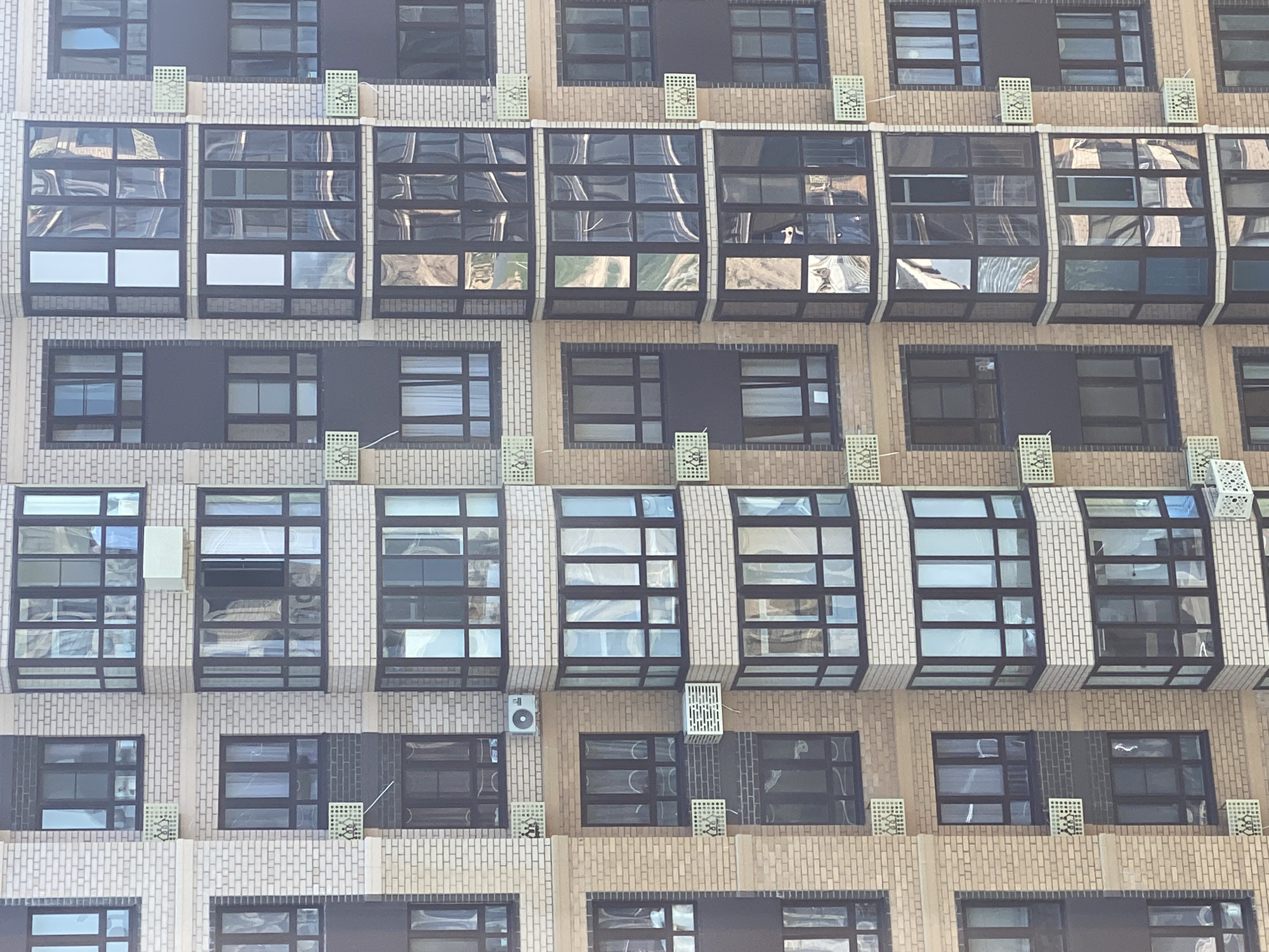 This is a close-up, eye-level, medium shot of a modern, multi-story apartment building. The building features a facade of beige and dark grey bricks, with numerous windows and balconies. Many of the windows are large and rectangular, with dark frames. Some of the windows are bay windows, extending outward from the building. Several balconies have glass enclosures.
The building is residential, as evidenced by the presence of air conditioning units mounted on the exterior walls. There are no people clearly visible in the image, though reflections in the windows suggest interior activity. The overall impression is one of urban development and contemporary architecture. The lighting suggests it is daytime, with sunlight casting shadows and highlighting the building's features.