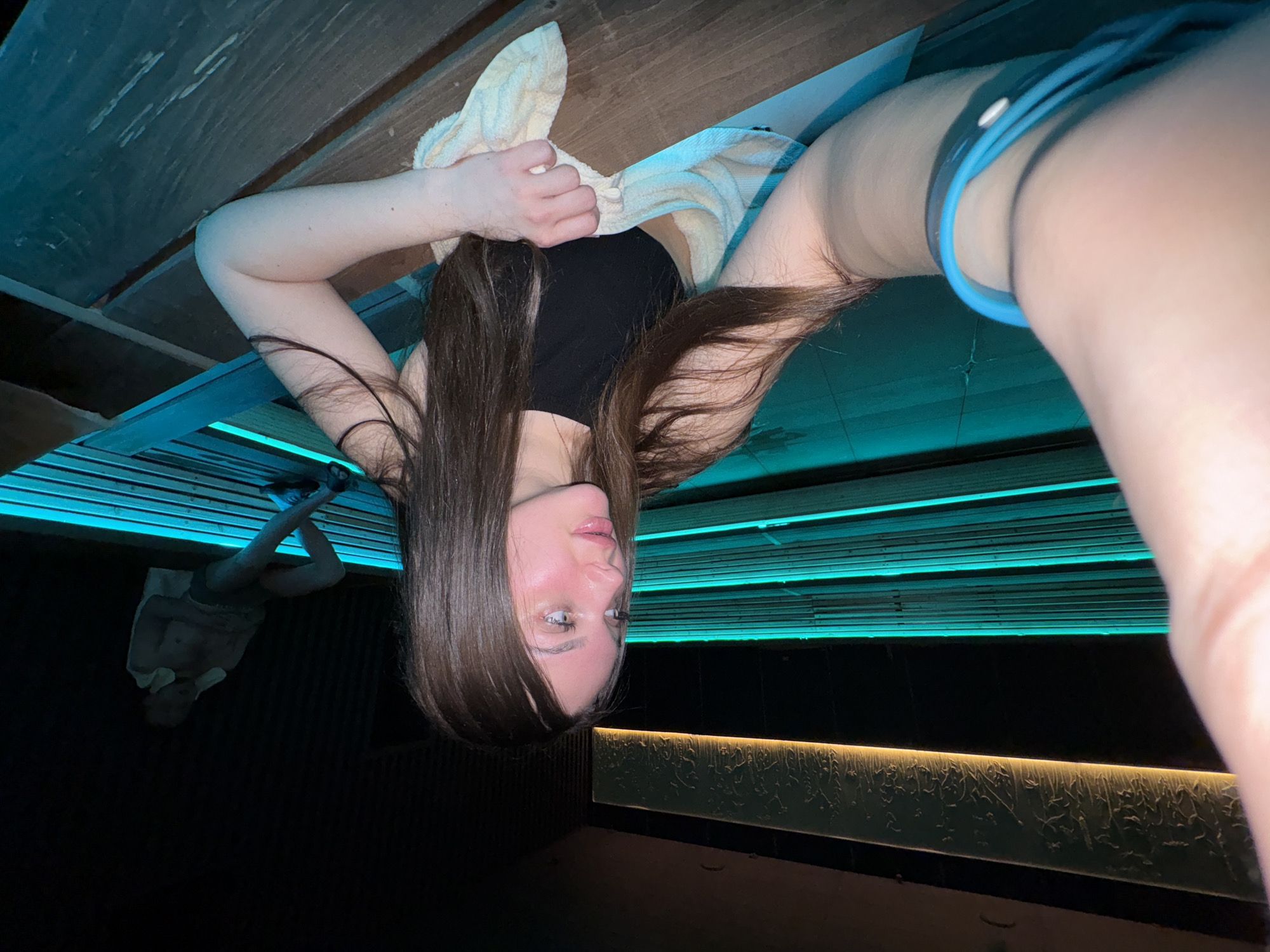 A woman lies in a tanning bed. She has long brown hair and a black tank top, and she is holding a towel. The tanning bed has blue lights. The location in the city of Yekaterinburg, Russia, is not specified.