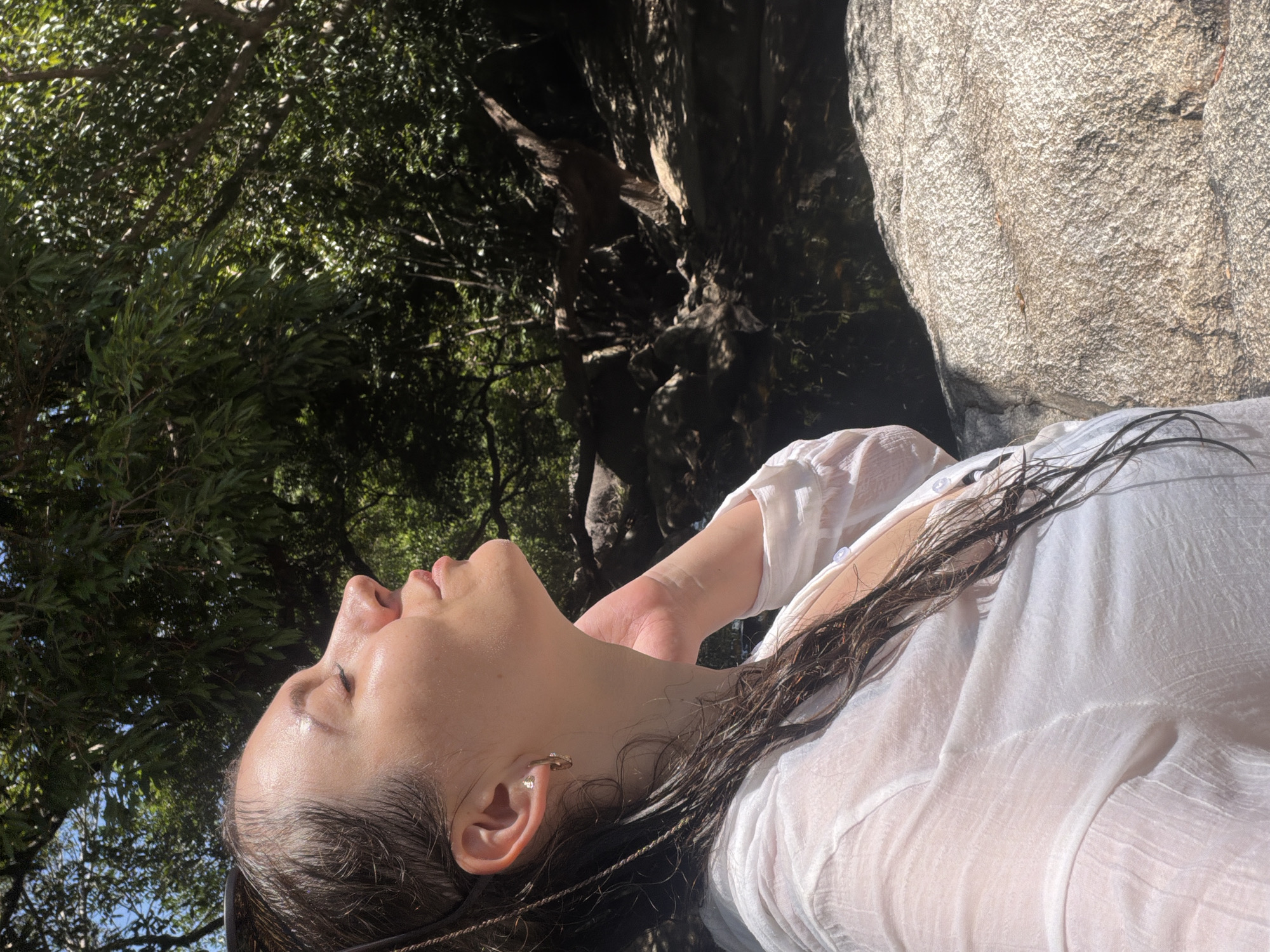 A medium shot captures a woman from the chest up, positioned on the right side of the frame, leaning back against a large, textured rock. She is facing left, with her head tilted back and eyes closed, basking in sunlight. Her wet, dark hair cascades down her shoulders, and she wears a sheer white shirt. A gold hoop earring adorns her left ear. Her right hand is raised, gently touching her neck. The background is a lush, dense forest with dappled sunlight filtering through the trees. A rocky stream bed is visible behind her, with dark water and more rocks. The overall scene suggests a natural, outdoor environment, possibly a waterfall or river area, within Hoa Thang, Vietnam.