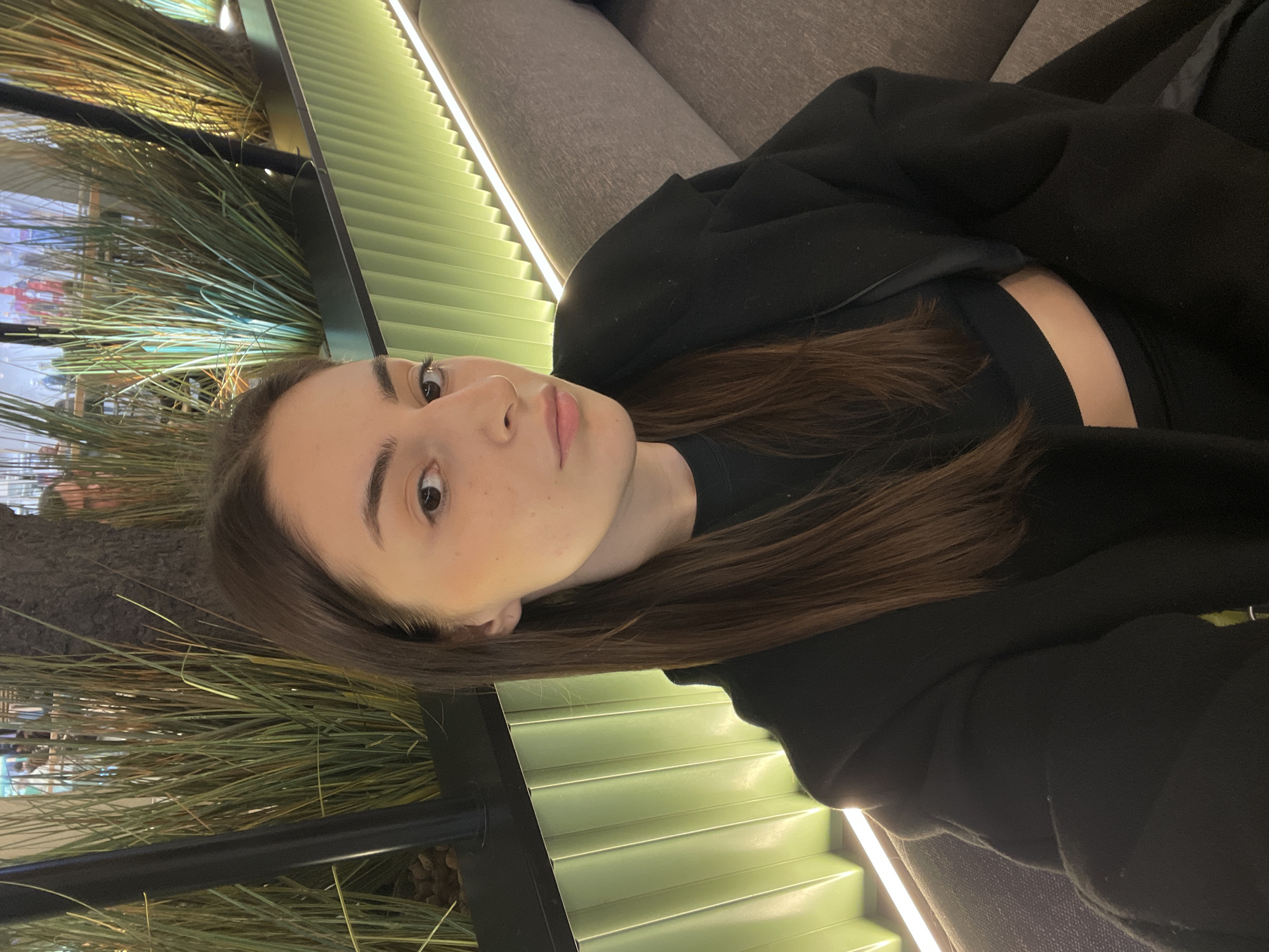 A young woman is seen in a close-up shot. She has fair skin, brown hair, and her face is angled toward the camera. She's wearing a black top and a black jacket or sweater. She appears to be seated or reclining on a gray upholstered surface. The background includes decorative reeds or grasses and a patterned wall with alternating light and darker green slats. The location is Moscow, Russia.