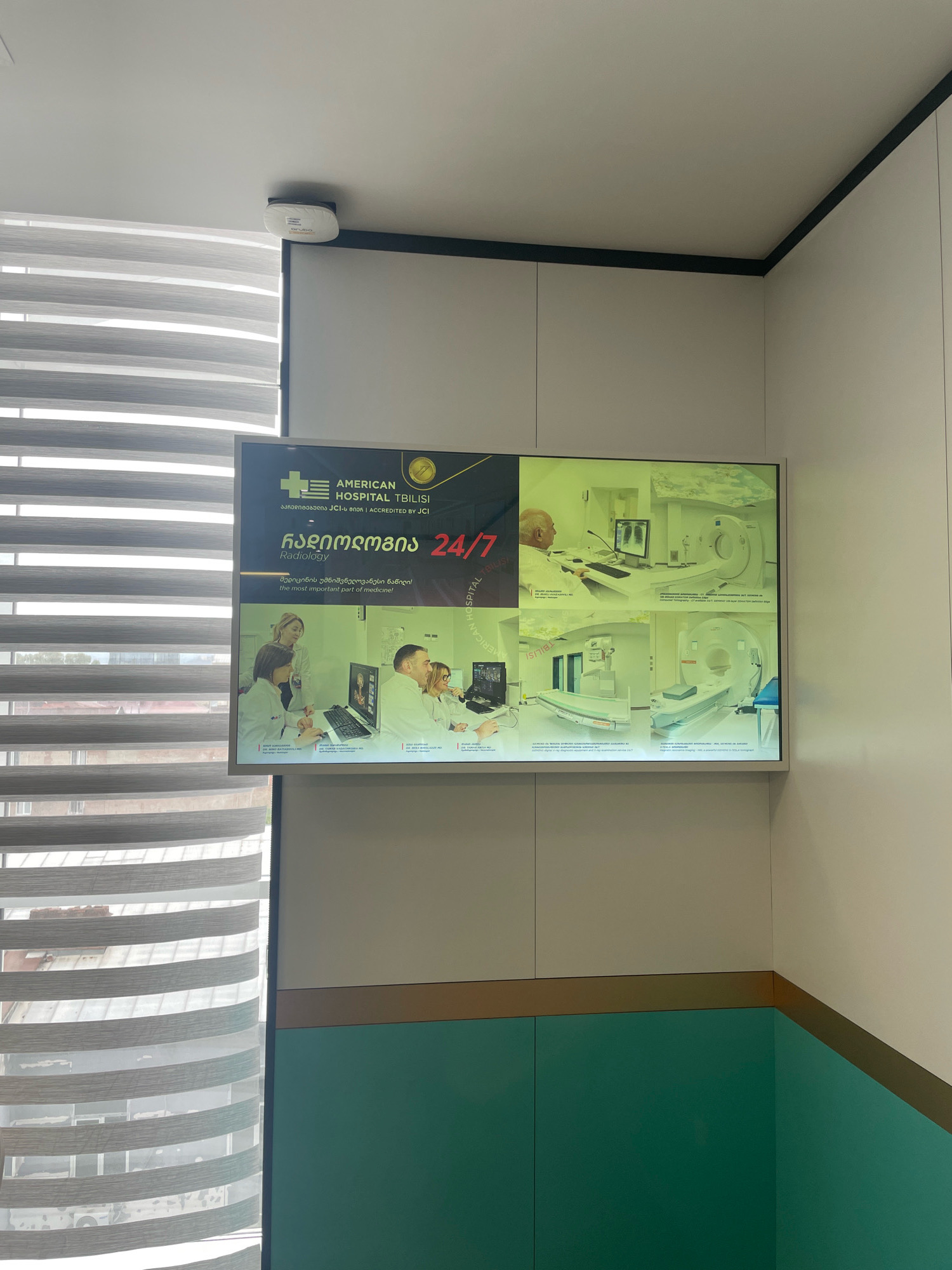 This image depicts an interior view of a medical facility, likely a hospital or clinic, in Tbilisi, Georgia. The central focus is a large digital display screen mounted on a wall. The screen prominently features the logo of "AMERICAN HOSPITAL TBILISI" and text in both Georgian and English, indicating that the facility is "ACCREDITED BY JCI." The displayed content highlights "Radiology" services, emphasizing that they are available "24/7." The screen also shows a collage of images related to medical imaging, including CT scanners and MRI machines, as well as several medical professionals reviewing images on computer monitors.
In the foreground, a window with horizontally slatted blinds is visible on the left side of the frame. These blinds are partially drawn, allowing natural light to filter in and revealing a glimpse of buildings and the urban environment outside, characteristic of Tbilisi. The wall behind the screen is painted in neutral tones, with a distinct horizontal band of darker color above a teal-colored section. A small white device, possibly a smoke detector or Wi-Fi access point, is mounted on the ceiling in the upper left corner of the image. The overall impression is one of a modern and well-equipped medical institution.