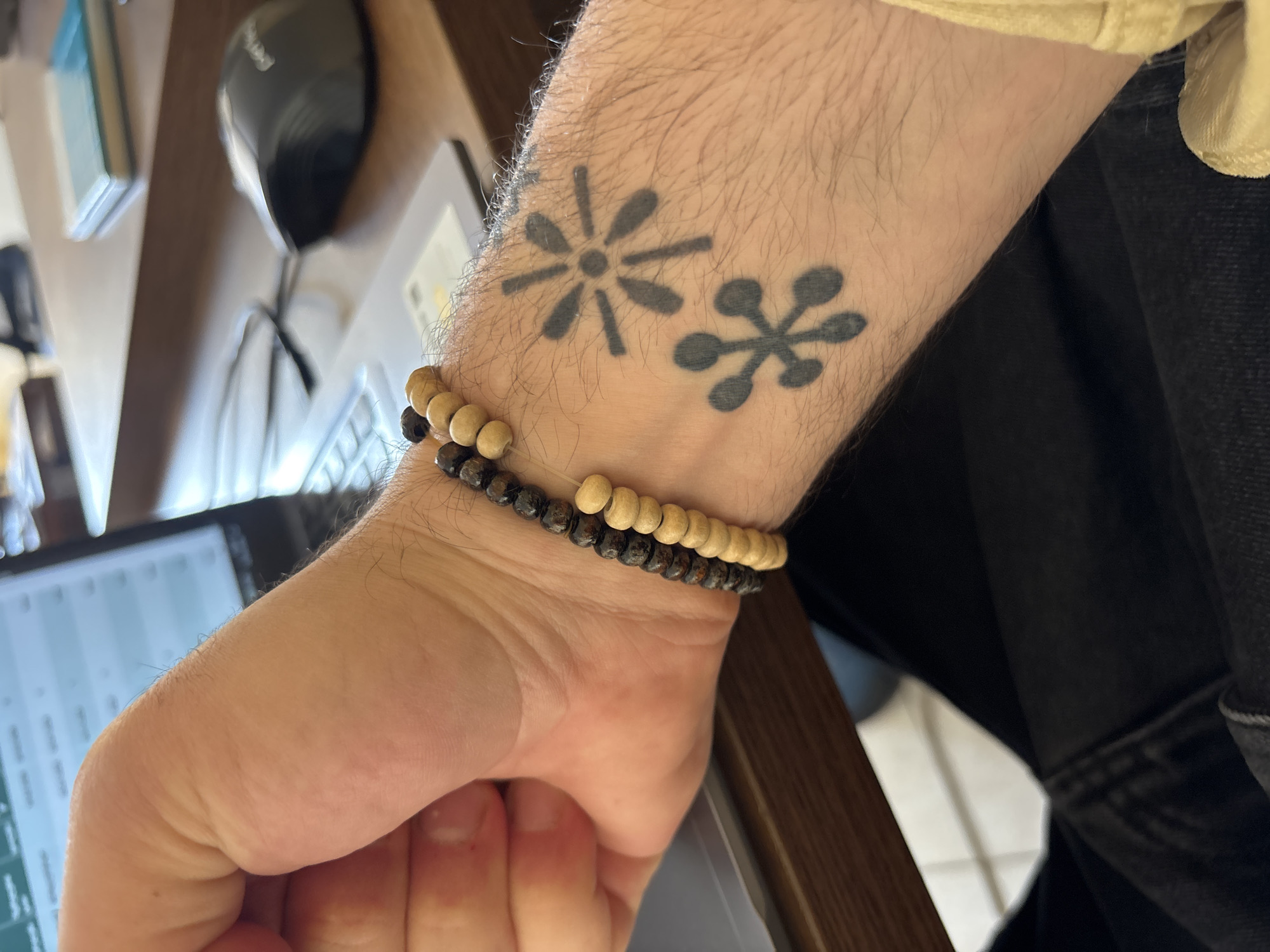 An indoor, close-up shot depicts a person's left wrist and forearm. The wrist is adorned with two beaded bracelets, one with light-colored wooden beads and the other with dark-colored beads. Above the bracelets on the forearm are two tattoos of asterisk-like shapes. The person's hand is partially visible, clutching what appears to be a laptop or tablet. The device's screen shows a grid of data. A wooden shelf or desk is in the background, supporting a black computer mouse. The clothing of the person is partially visible, suggesting a light-colored shirt and dark-colored jeans or pants. The location appears to be indoors, with a glimpse of a tiled floor in the periphery. There's no specific information to suggest this is in Tbilisi, Georgia.