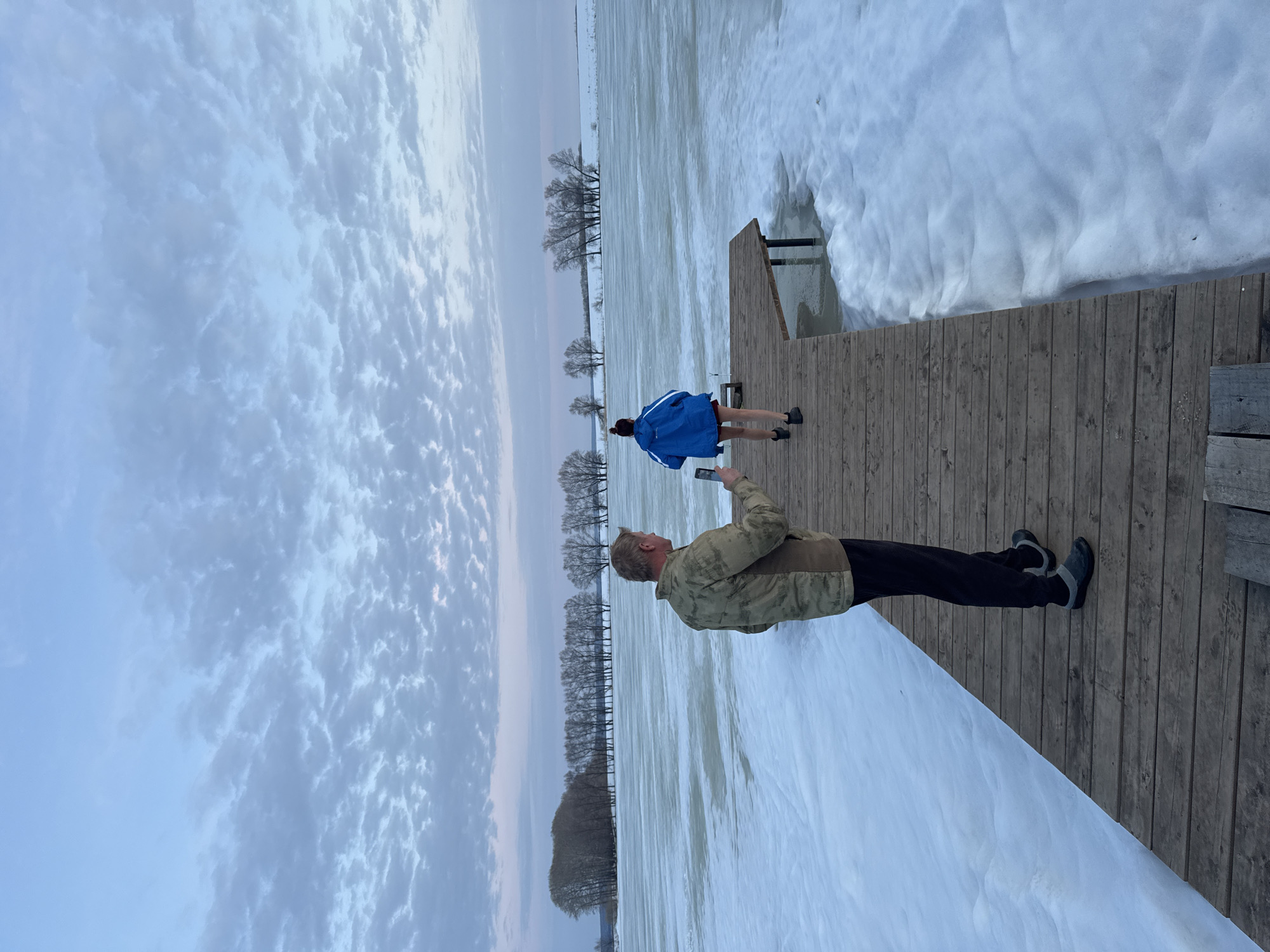 An outdoor scene depicts a partially frozen body of water with a wooden dock under an overcast sky. The location is Unknown, Unknown. An adult male, wearing a camouflage jacket, dark trousers, and dark shoes, stands on the dock in the foreground, facing away from the camera. He holds a smartphone in his right hand, directed towards an adult female ahead of him. The female, wearing a blue hooded jacket, dark shorts, and dark footwear, walks along the dock towards its end, away from the camera. The water surrounding the dock is covered in slushy ice and snow, with areas of open water visible further out. In the background, a distant shoreline features a line of bare deciduous trees and a snow-covered landscape under the cloudy sky.