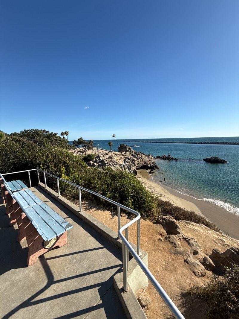 The image is a scenic view from a coastal area in Newport Beach, United States, featuring a clear blue sky and a portion of the Pacific Ocean. In the foreground, there is a light blue bench situated on a concrete walkway, protected by a silver-colored railing. The walkway leads to a natural overlook, with green shrubbery covering the adjacent land. The beach is visible below, with gentle waves lapping the sandy shore. Further out, a breakwater structure stretches into the ocean, defining a harbor area. The lighting suggests a sunny day, with strong shadows cast by the railing and bench.