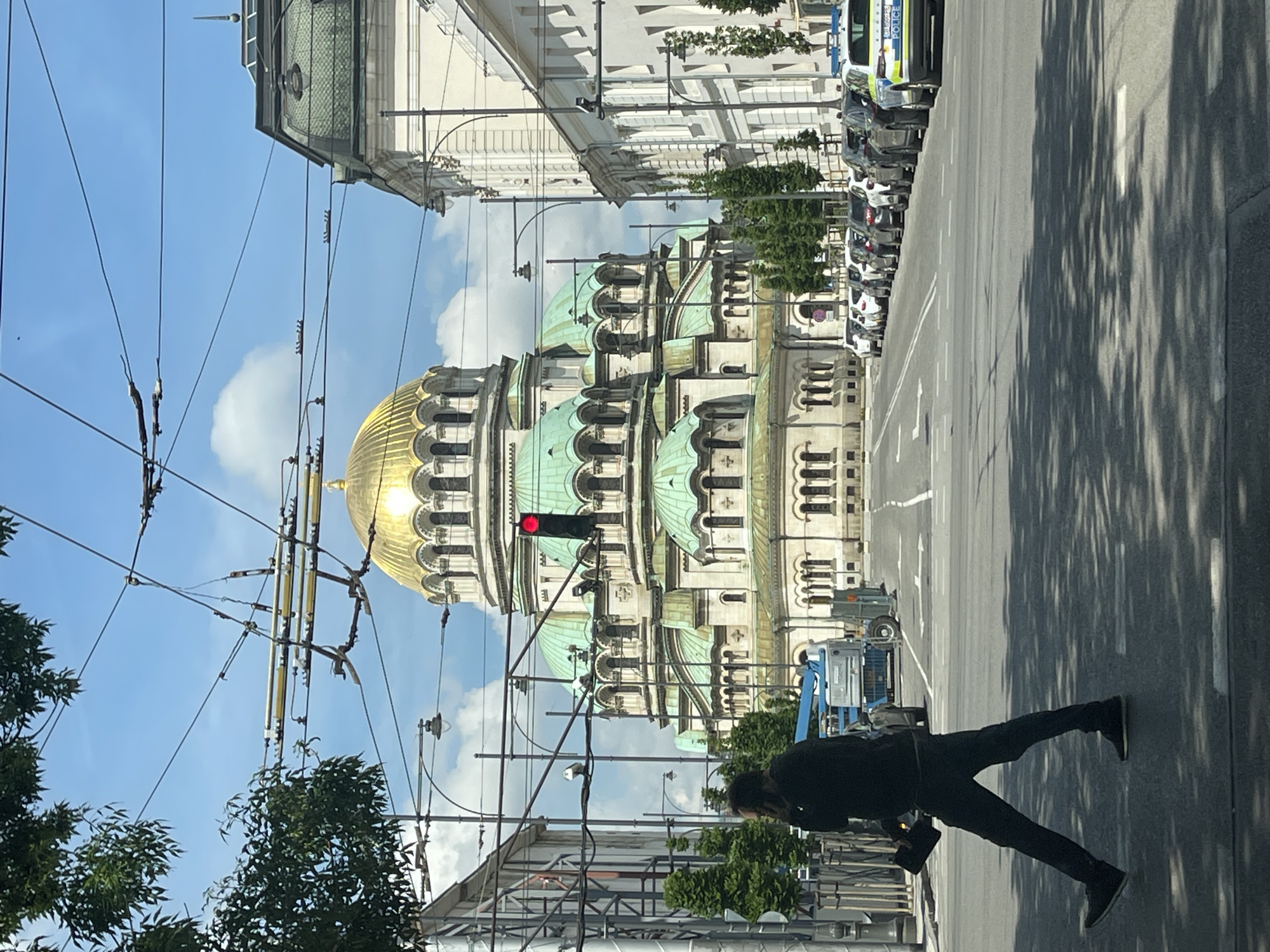 An eye-level outdoor daytime shot depicts a city street in Sofia, Bulgaria, featuring the Alexander Nevsky Cathedral as a prominent element. A person dressed in black stands in the foreground, partially obscured by trees and utility lines. In the background, a line of vehicles, including police cars, is parked along the curb. The Cathedral’s distinctive golden dome and green tiers stand out against a partly cloudy blue sky. A red traffic light is visible near the Cathedral. The city street appears wide, with building facades lining the sides and tree shadows stretching across the pavement.