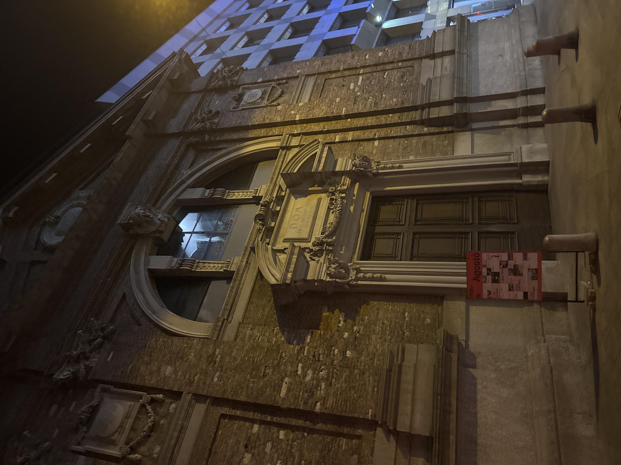 The image shows the exterior of an old building at night. The building is made of brick and has classical architectural elements, including an arched window and ornate carvings above a large wooden door. A sign with the word "Agosto" (August in Italian) is posted on an easel to the left of the entrance. In the background, a modern building with blue lighting is visible. The street is empty except for several bollards. The scene is illuminated by artificial light, casting long shadows. The location is not identifiable as Lugano, Switzerland from the visual information present.