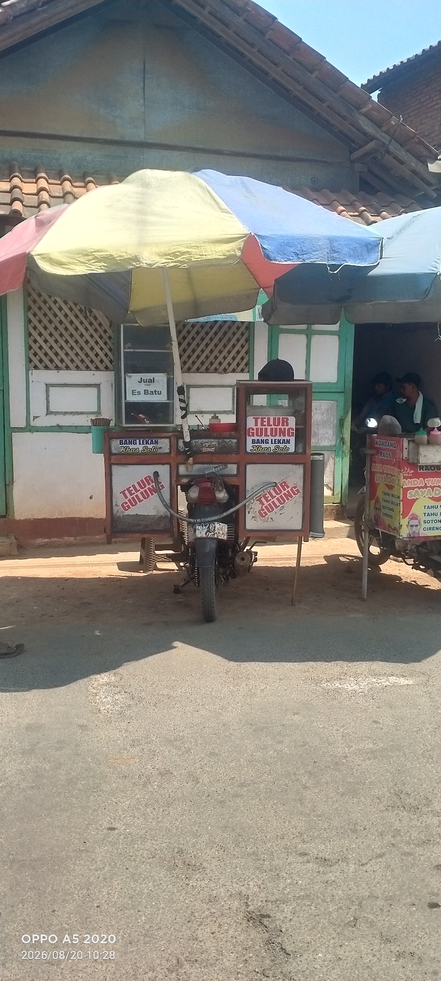 A street vendor's cart, possibly in Bugel, Indonesia, is set up for business under a multi-colored umbrella. The cart is a metal frame with a motorcycle engine and wheels, suggesting it is mobile. On the cart, signs advertise "Telur Gulung" (rolled egg), with one sign specifying "Bang Lekan Khas Solo," indicating a specialty from Solo. Another smaller sign on the cart reads "Jual Es Batu" (selling ice blocks). To the right of the cart, another vendor is visible inside a shop, with signs for "Kondang Raos" and various food items including "Tahu Malia," "Tahu Bulat," and "Sotong." The environment appears to be a sunny day with a clear blue sky. The ground in front of the cart is a paved road, and a shadow is cast by the cart and umbrella. A motorcycle is attached to the back of the cart.