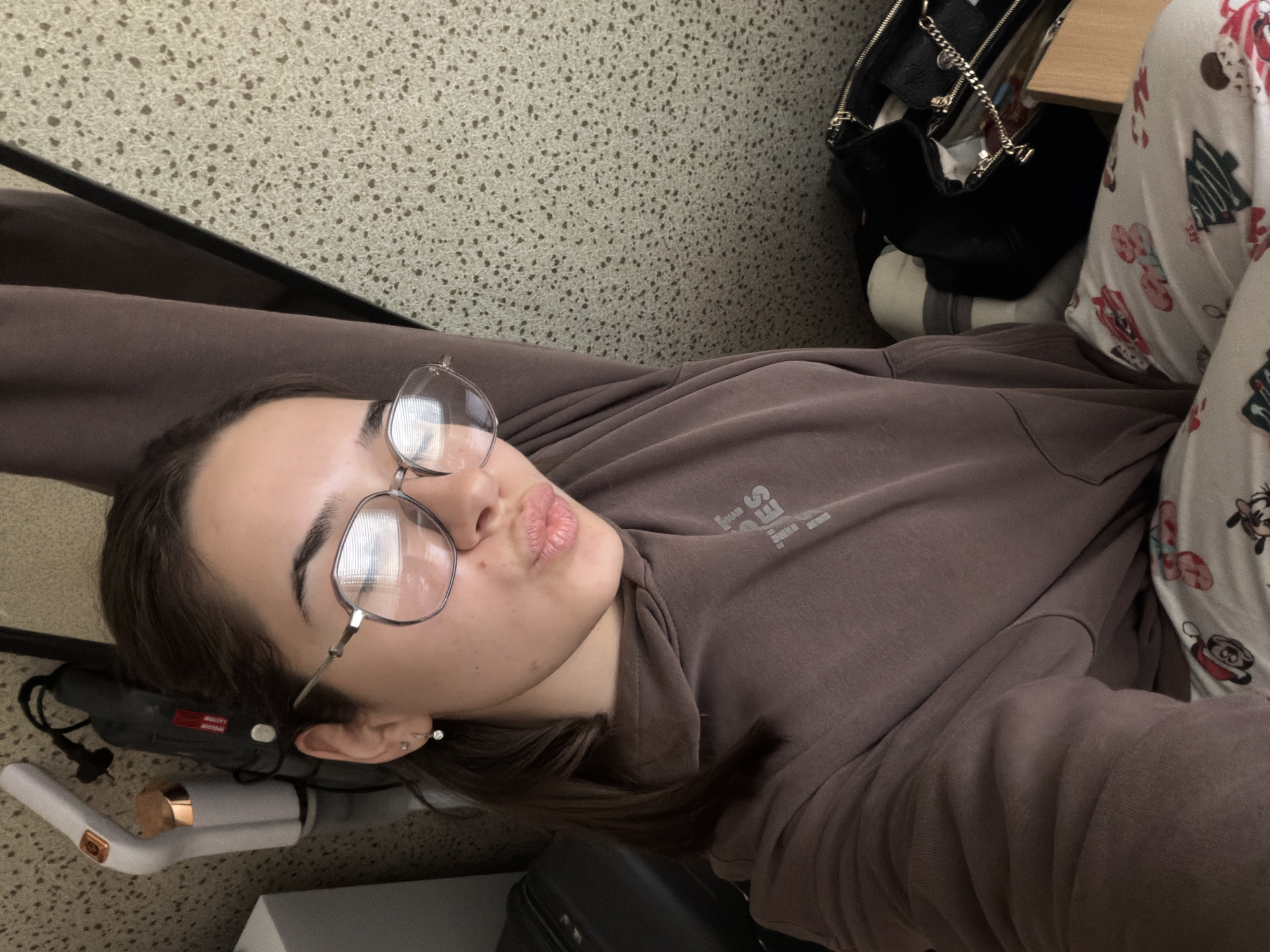 An angled, close-up selfie shows a young woman lying down in an indoor setting. She is looking towards the camera with a playful "duck face" expression. She has dark hair and wears rimmed eyeglasses. A small earring is visible on her left ear.
She is dressed in a casual brown or mauve hooded sweatshirt, featuring some partially visible, mirrored text on the chest, possibly reading "ESSENTIALS." What appears to be matching sweatpants are also visible. On her right side, white pajama bottoms with cartoon character prints, including Mickey Mouse, Minnie Mouse, Goofy, and Christmas trees, are partially seen.
The background consists of a light-colored, speckled textured surface, likely a wall or floor. To the right, a black handbag with a metallic chain strap rests beside a light wooden surface. Below her head, a white and rose gold hair styling tool, resembling a curling iron, is visible with its black cord.
The image conveys a relaxed and casual mood indoors. No specific time of day or weather conditions can be determined from the photo.
