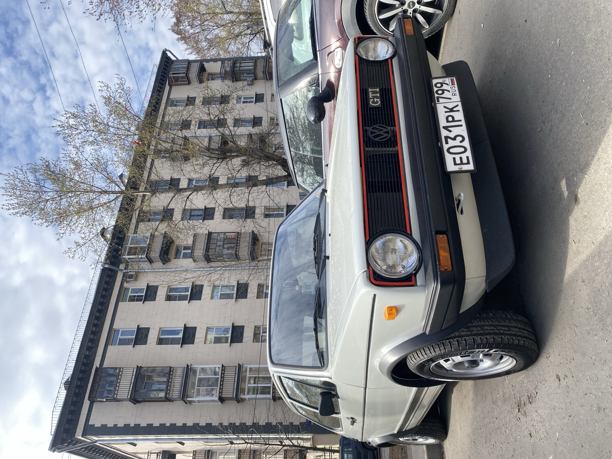 A slightly tilted eye-level shot captures the front of a white Volkswagen Golf GTI with the license plate "E031PK 799 RUS," parked on a city street in Khimki, Russia. The car's red-trimmed black grill, round headlight, and a glimpse of the front tire are prominent features. The GTI is parked alongside a reddish-brown vehicle. In the background, an aged apartment building rises, its facade featuring multiple windows and ornate balconies, partially obscured by the bare branches of a tree. The sky overhead is partly cloudy. The setting suggests an urban residential area.