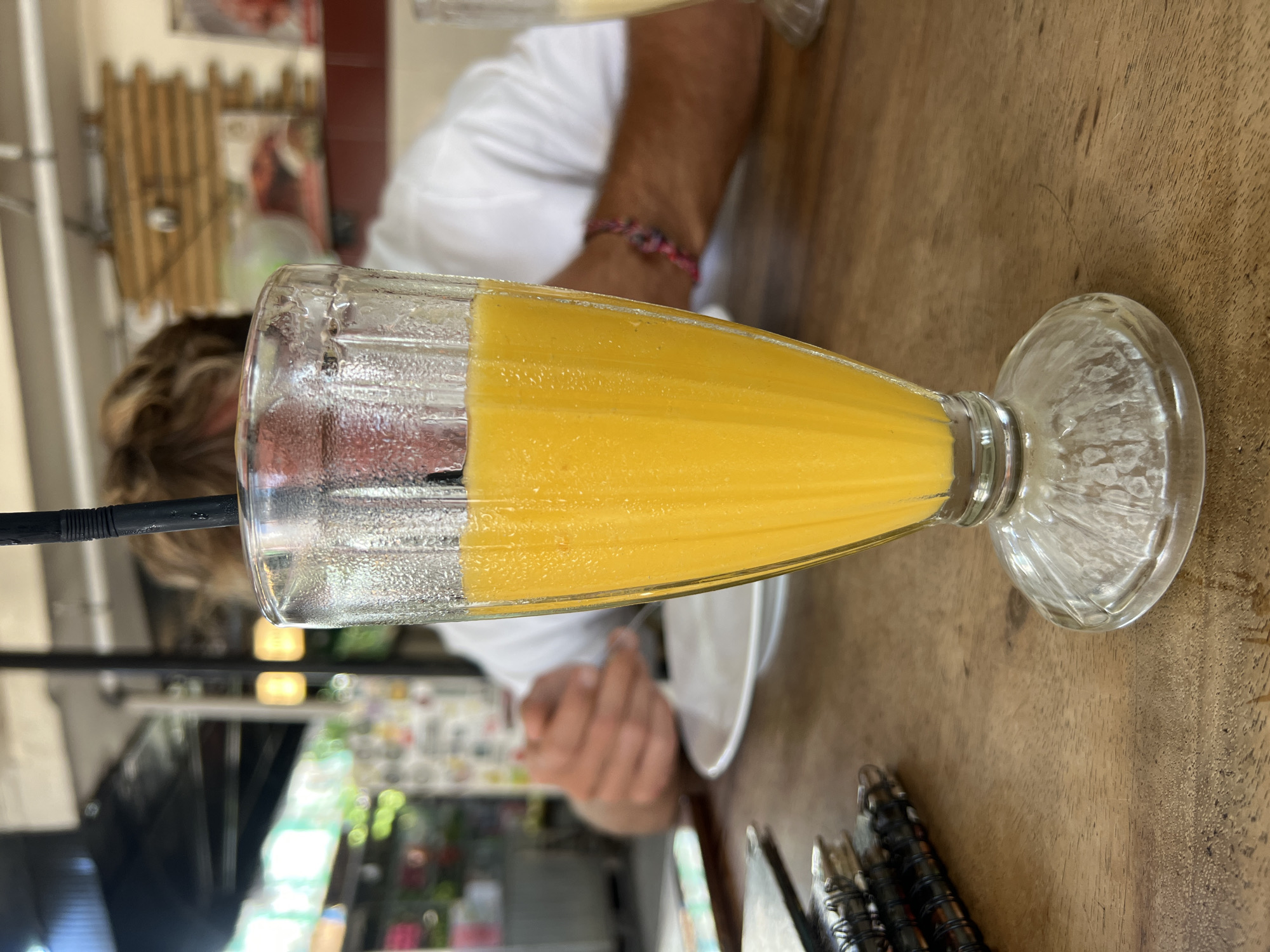 A close-up, eye-level shot shows a tall, fluted glass filled with a vibrant orange beverage, likely a smoothie or juice, sitting on a rustic wooden table. The glass is covered in condensation. A black straw extends from the drink. In the background, a person with blondish hair, wearing a white t-shirt and a colorful braided bracelet on their wrist, is partially visible, their hands blurred as they appear to be eating. The setting appears to be an outdoor cafe or restaurant in Kuta, Indonesia, suggested by the blurred elements of a covered outdoor seating area, ambient lighting, and foliage in the distance. A stack of notebooks or menus is visible to the left of the glass.