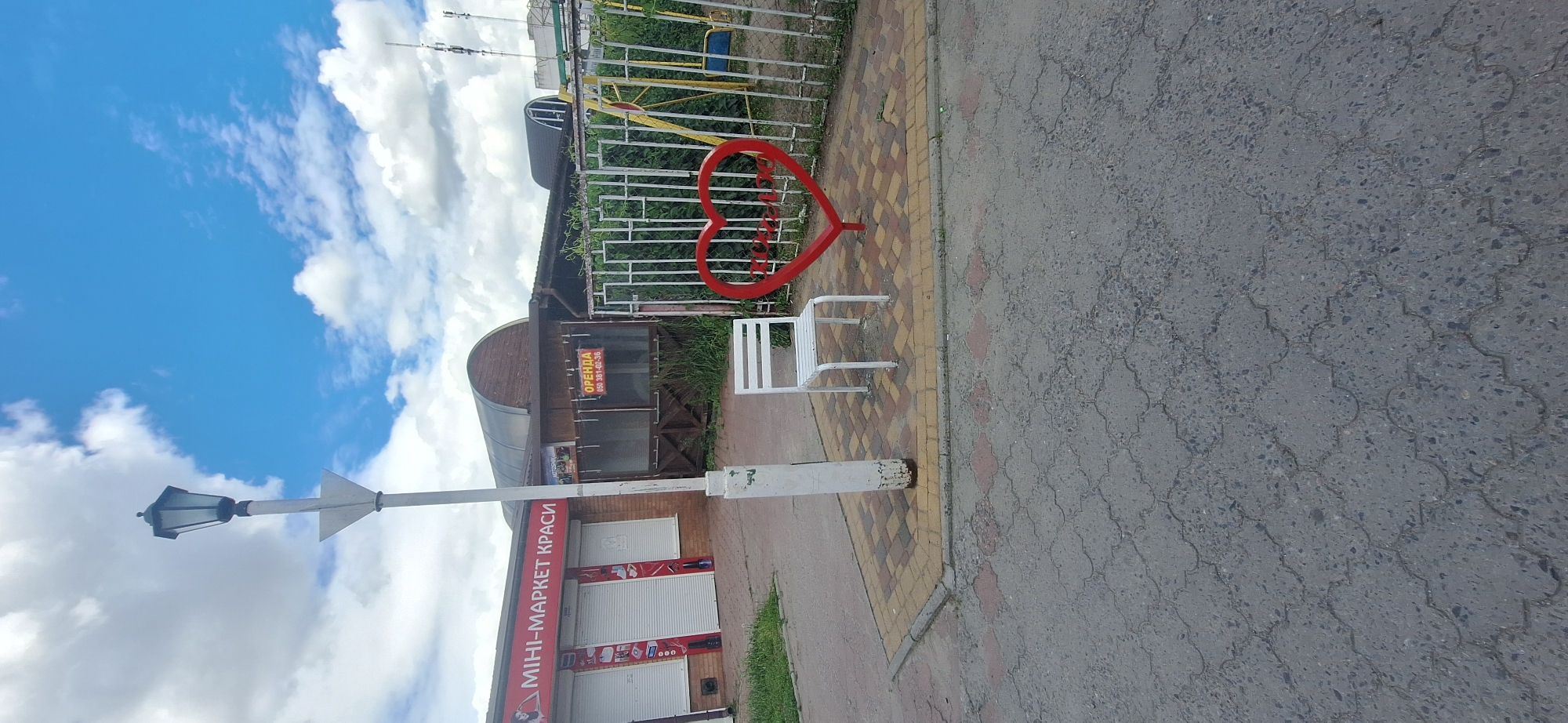 The image shows a street scene in Bila Tserkva, Ukraine, under a cloudy blue sky. In the foreground, asphalt paving stones with a distinctive pattern are visible. A short distance further, a paved area with rectangular and square interlocking bricks forms a pathway. A white metal chair is placed on this paved area, next to a white lamppost. Behind the chair, a large red heart-shaped sculpture is positioned in front of a metal fence. The fence encloses an area with some vegetation and a swing set. To the left of the lamppost is a building with a red sign that reads "МІНІ-МАРКЕТ КРАСИ" (MINI-MARKET BEAUTY). This building has closed white shutters. Further to the right is a smaller, darker building with a sign that says "ОРЕНДА" (RENT) with a phone number. The overall scene suggests an outdoor commercial area.