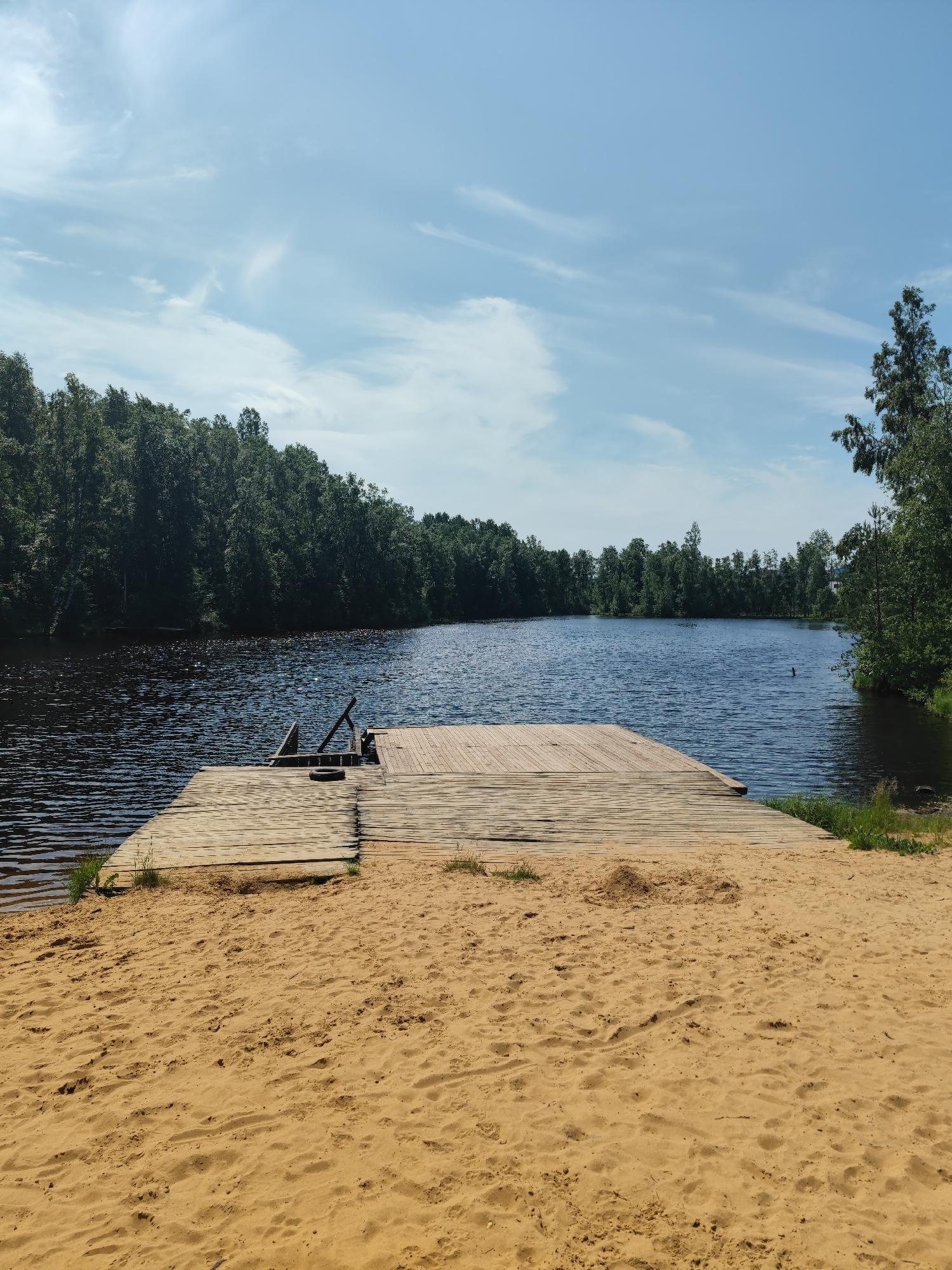A wooden dock extends from a sandy beach into a dark, calm lake. The lake is surrounded by a dense forest of green trees under a blue sky with scattered white clouds. The scene depicts a serene natural landscape.