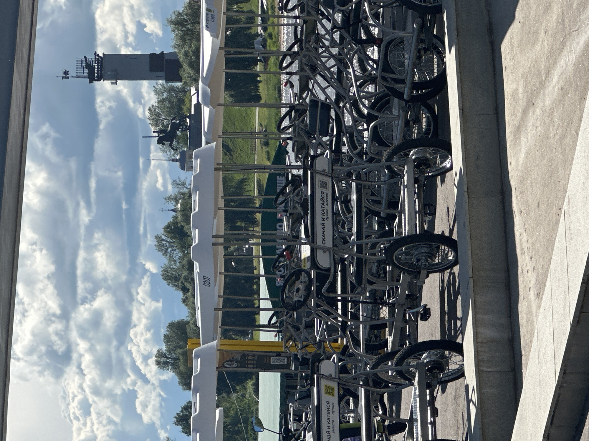 The image shows a bike rental station with numerous bicycles parked in racks. The setting appears to be outdoors, possibly near a body of water, with trees and a cloudy sky visible in the background. A tall tower structure is also visible in the distance. The bicycles are arranged in multiple tiers, suggesting a large capacity rental service. Some of the bicycles have visible markings, including numbers and text. The scene is brightly lit, indicating it is daytime, and the presence of clouds suggests a partly cloudy day. The overall impression is that of a public amenity available for recreational use. The visible text includes "СКАЧАЙ И КАТАЙСЯ" which translates to "Download and Ride" and "вместе лучше" meaning "together is better", suggesting a modern, app-based rental system. Numbers "0307" and "0300" are also present on some of the units. The context of Veliky Novgorod, Russia, implies this is a common sight in an urban or park setting within that city. There are no people clearly visible in the frame, and the focus is on the rental infrastructure and the bicycles.