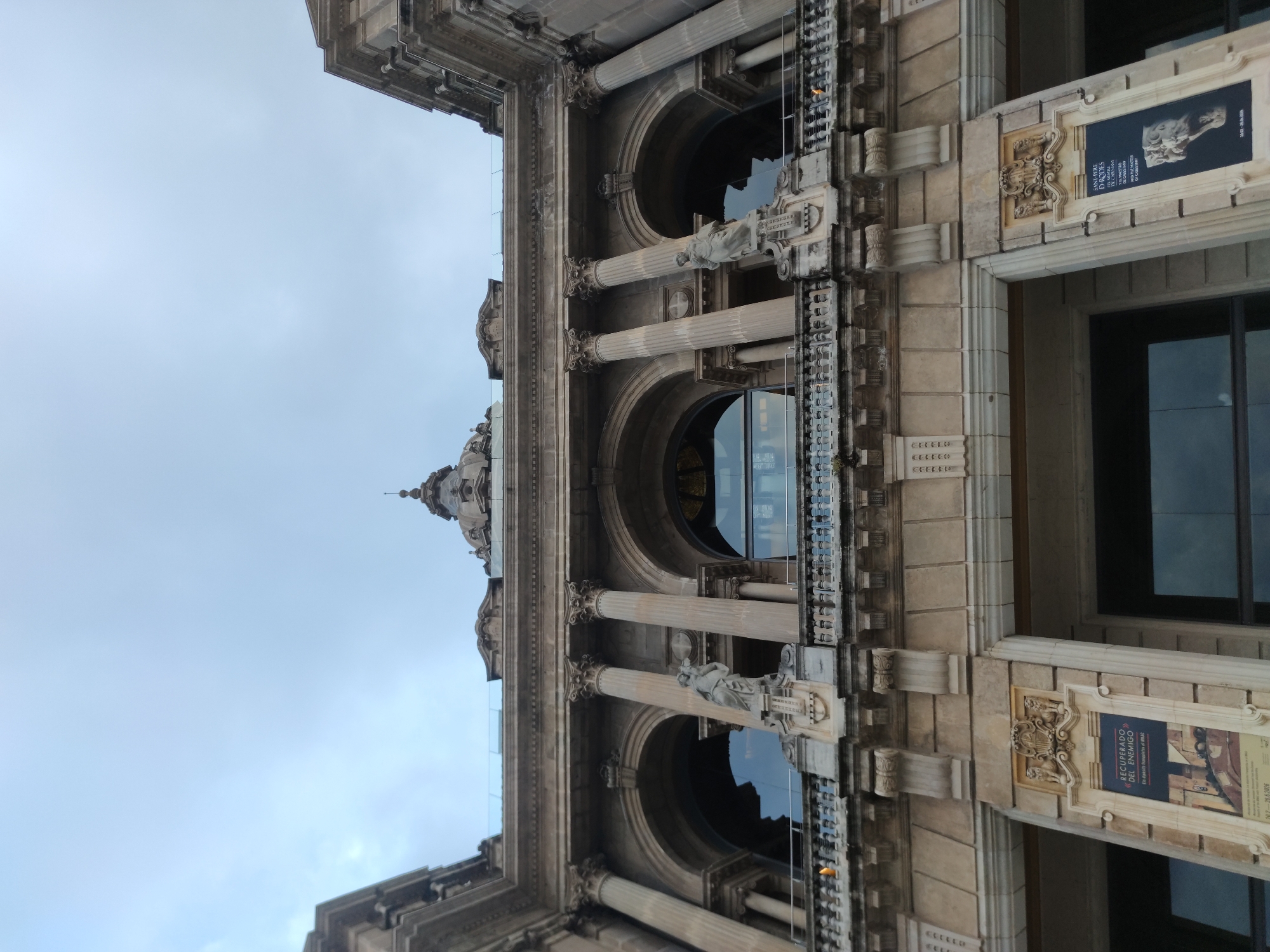 The image shows a low-angle view of a classical building in Barcelona, Spain. The building is constructed from tan-colored stone, with prominent architectural features such as columns, arches, statues, and ornate carvings. A central dome is visible at the top of the building. Banners advertising cultural exhibits are displayed along the facade. The sky is overcast, suggesting a cloudy day.
