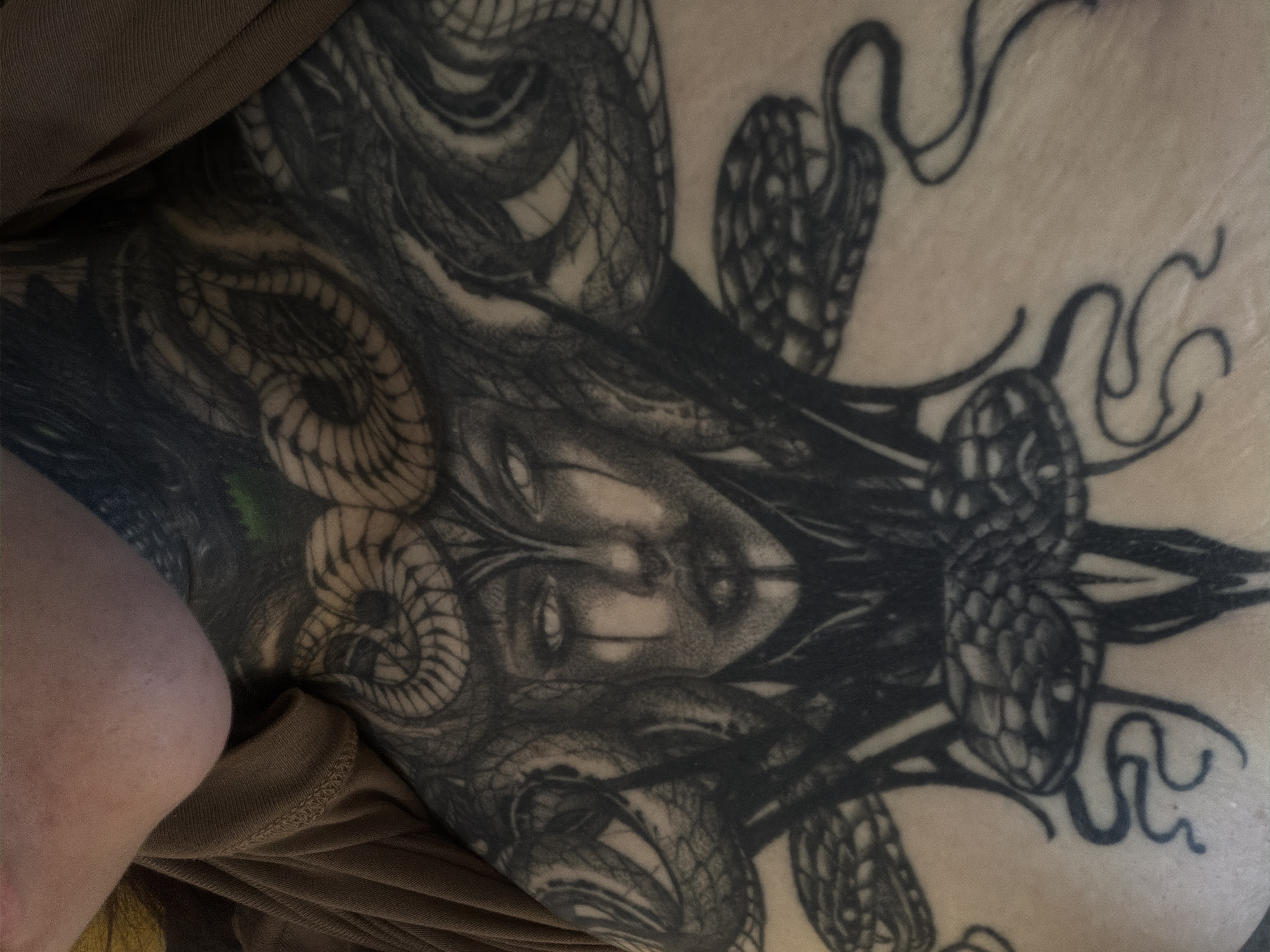 The image displays a close-up, downward-angled view of a large, monochrome tattoo on light-toned human skin. The tattoo depicts a stylized Medusa head, centrally featuring a female face with prominent white, glowing eyes from which dark, tear-like markings descend. Snakes form the hair, with additional serpents integrated into the design above and below the central face, rendered with intricate scales and shading. A segment of brown, ribbed fabric, consistent with clothing, is visible along the upper left and right edges of the frame. The precise location within the city of Chaikovsky, Russia, cannot be determined from the visual information provided in the image.