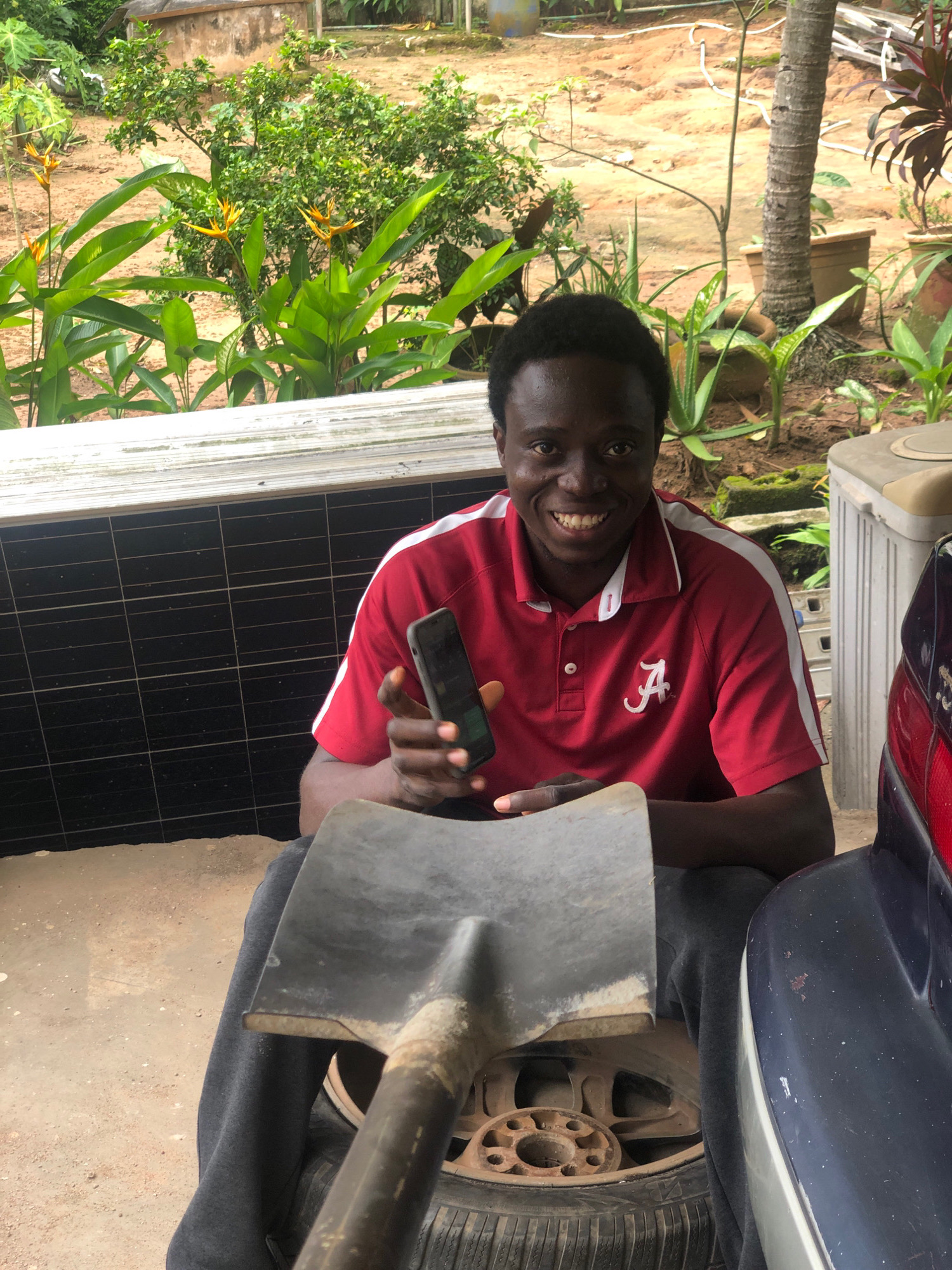 A dark-skinned man with short, black hair and a wide smile is seated outdoors, holding a smartphone in his right hand. He is wearing a red polo shirt with white trim on the collar and sleeves, and an "A" logo is visible on the left chest. The man is sitting on a car tire, which is placed on the ground. In front of him, a shovel is resting on the tire. The background features lush green vegetation, a palm tree, and a concrete structure. The ground appears to be sandy and dirt. The scene is likely in Iju, Nigeria, given the environmental context.
