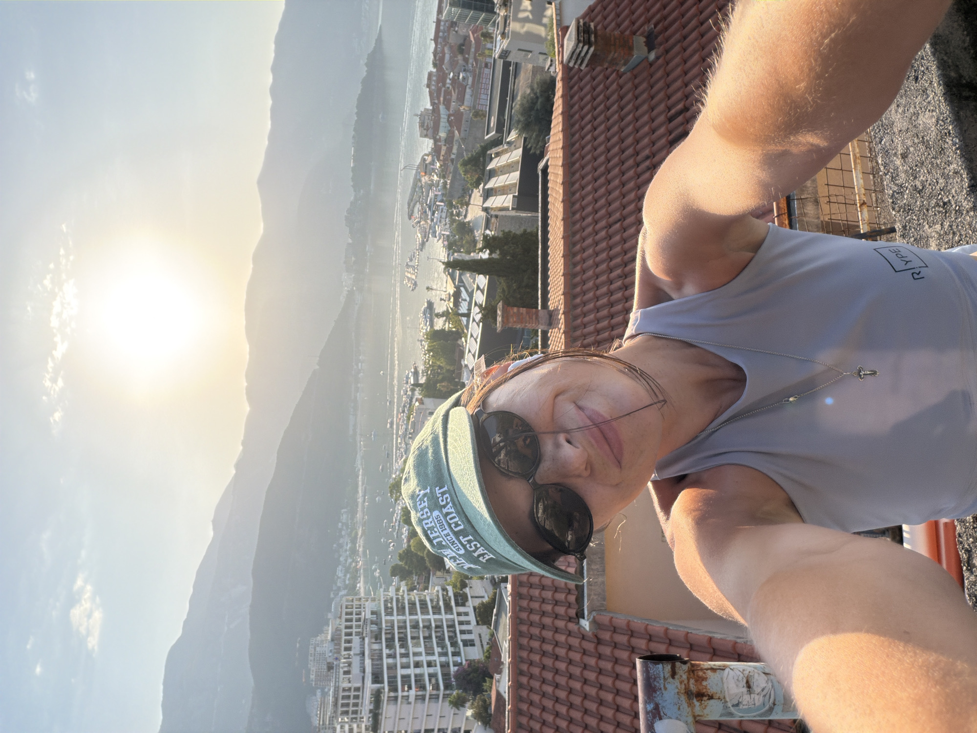 A woman with dark hair, wearing a light gray tank top and a green baseball cap, takes a selfie on a rooftop overlooking the city of Budva, Montenegro. She is wearing sunglasses and a necklace with a cross pendant. The background shows a view of the city, a harbor with boats, and mountains in the distance. The sun is setting, casting a warm glow over the scene. The woman is smiling and looking at the camera. She appears to be standing on a rooftop with red tiles.