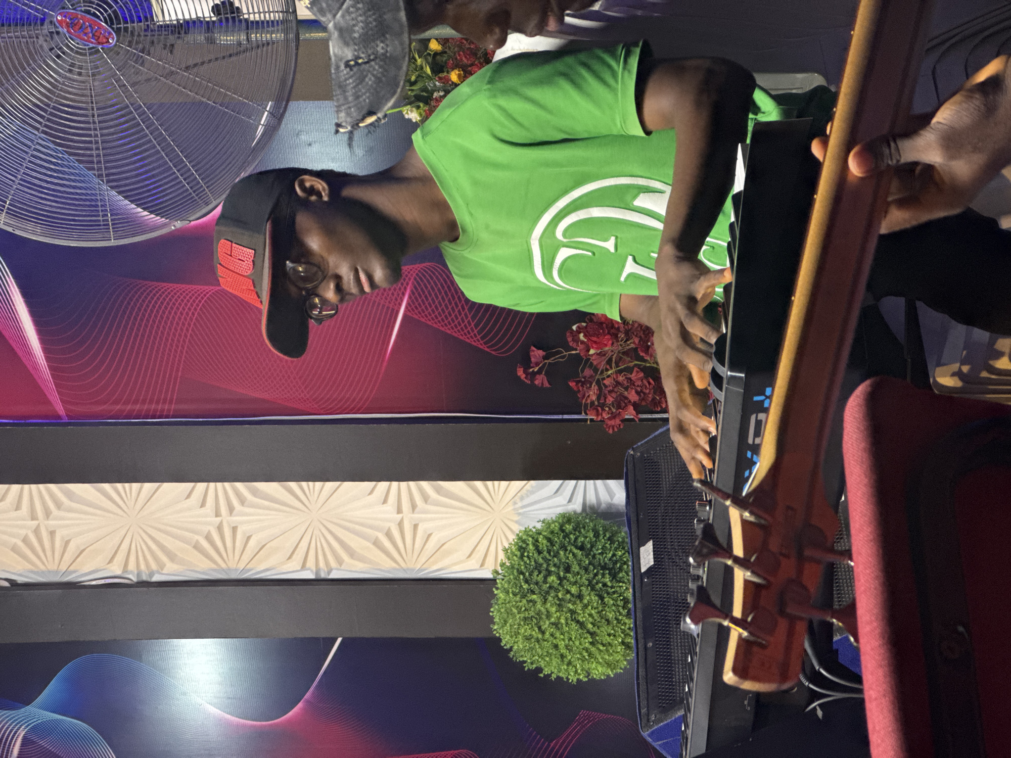 A medium shot captures a musician wearing a green t-shirt and a black baseball cap playing a keyboard. His hands are positioned on the keys, suggesting he is performing. To the right, a person is holding a stringed instrument, possibly a bass guitar. The backdrop is a patterned wall with a combination of geometric and wavy designs, colored in hues of red, blue, and white. A potted plant and a decorative floral arrangement add to the scene's ambiance. The overall setting appears to be a stage or performance area, potentially within the city of Onitsha, Nigeria. A large fan is visible in the upper left corner of the image.