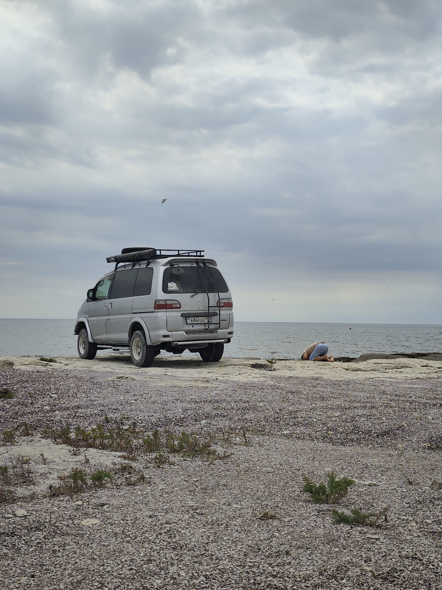 An overcast day at the coast of Aqshuqyr, Kazakhstan. A silver minivan is parked on a rocky, shell-strewn beach near the water's edge. A person in light blue shorts and a tan shirt is kneeling on the ground facing away from the camera, with their head bowed towards the earth. The sea is calm and stretches to the horizon. Birds are visible in the sky.