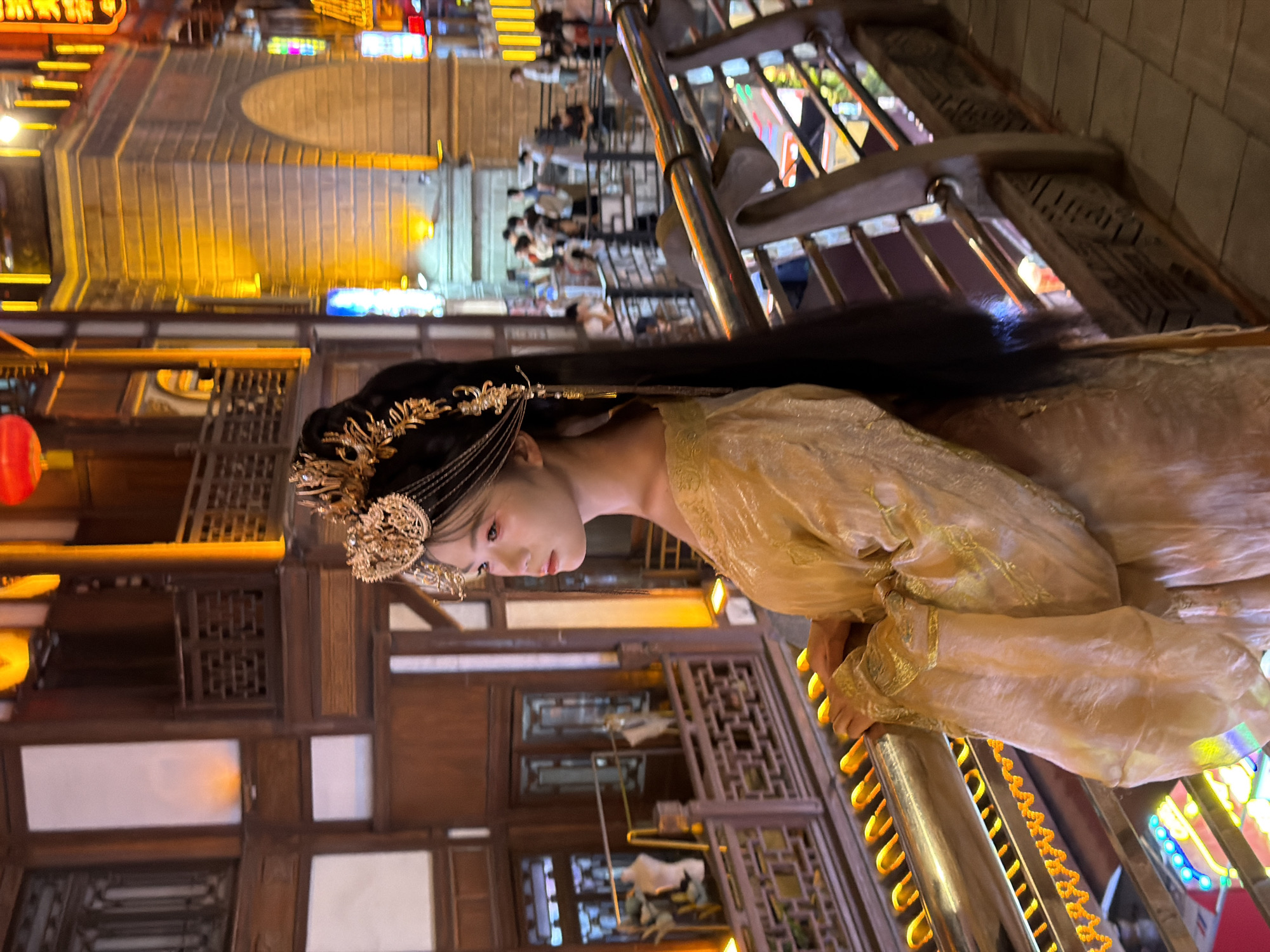 A young East Asian woman, with dark hair styled elaborately with a golden headdress, stands on a balcony or walkway overlooking a street scene at night in Yuzhong District, China. She is wearing a traditional golden gown with flowing sleeves and intricate gold embroidery. Her gaze is directed to her left, away from the camera, with her hands resting on a metal railing. The background features traditional Chinese architecture with wooden balconies and illuminated brick buildings. The street below is bustling with people, and colorful neon lights are visible, suggesting a commercial or entertainment area. The lighting in the scene is warm and atmospheric, with golden hues dominating from streetlights and building illuminations.