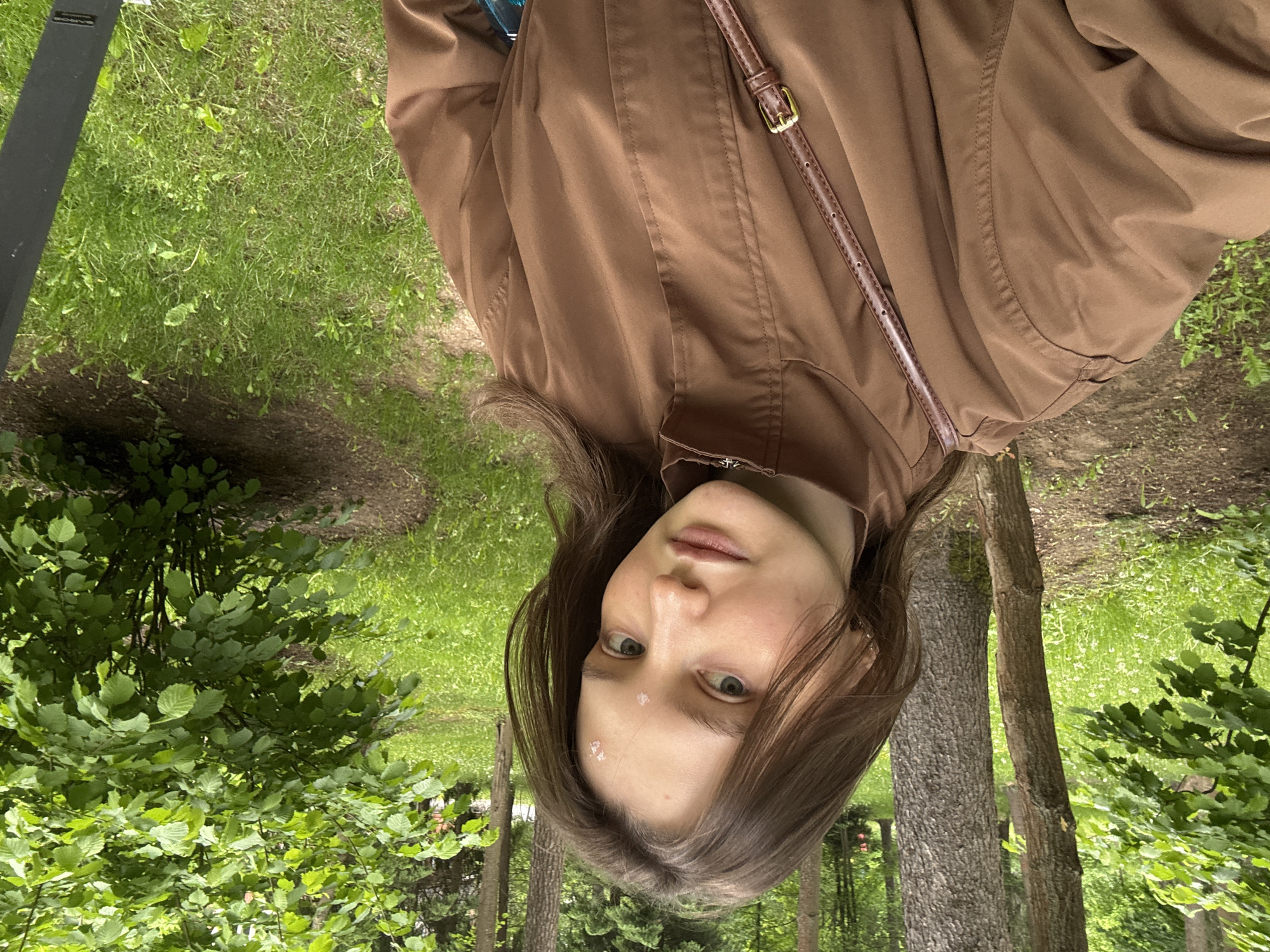 A young woman is facing the camera, her head and shoulders positioned in the lower center of the frame. She has fair skin, dark hair, and light-colored eyes. A few small white objects are stuck to her forehead. She is wearing a brown jacket with a thin brown strap across her torso.
The background features a park-like setting with green grass, trees, and foliage. Tree trunks are visible behind the woman, suggesting she is standing in a wooded area. The location is Vnukovo, Russia.