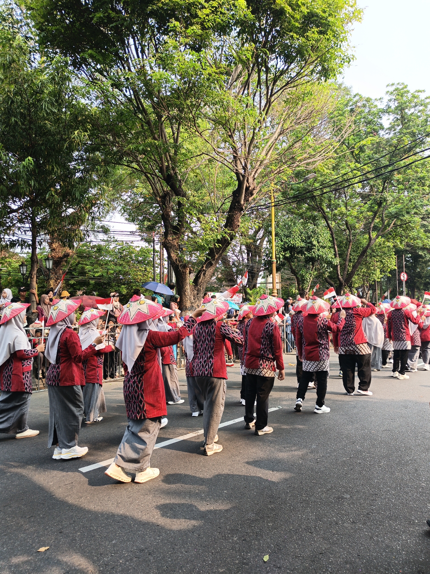 The image captures a cultural parade on a street in Gayamprit, Indonesia. A group of individuals, primarily women dressed in traditional attire, are participating. They are wearing red and white patterned tops and grey skirts, complemented by distinctive conical hats adorned with a starburst design. Some are holding small Indonesian flags. The procession is taking place on an asphalt road, lined with trees and streetlights. Spectators are visible in the background, some holding umbrellas. The scene appears to be illuminated by natural sunlight, casting shadows on the road.
