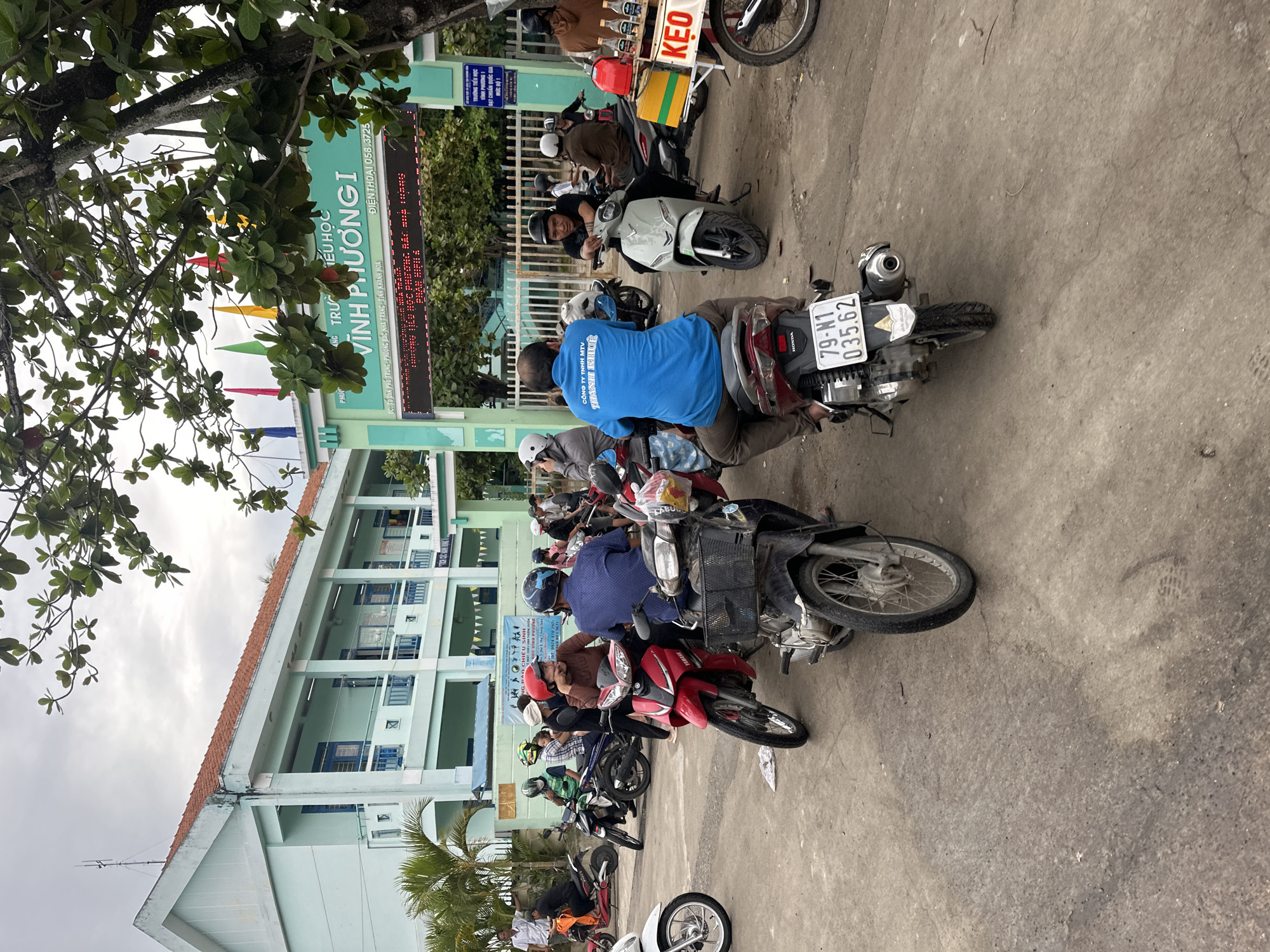 This is a wide-angle, eye-level, outdoor shot taken on a cloudy day in Nha Trang, Vietnam, likely in front of a school. The foreground is dominated by a paved area with several motorcycles parked and some people on them. One man in a bright blue shirt is on a scooter with a license plate reading "79-N1 035.62". Another scooter in front of him has a basket with items, including what appears to be a "KEO" sign, suggesting a vendor. To the right, a person in a brown shirt is on a motorcycle with a rack holding bottles. In the midground, more people and motorcycles are visible. Behind the motorcycles, a green school building with a red-tiled roof is prominent. Above the entrance to the school, a large digital sign displays text in Vietnamese. The school appears to be "TRƯỜNG TIỂU HỌC VĨNH PHƯỚNG I". A tree with green leaves partially obscures the top of the image. The overall scene depicts a busy entrance area, possibly during school drop-off or pick-up.