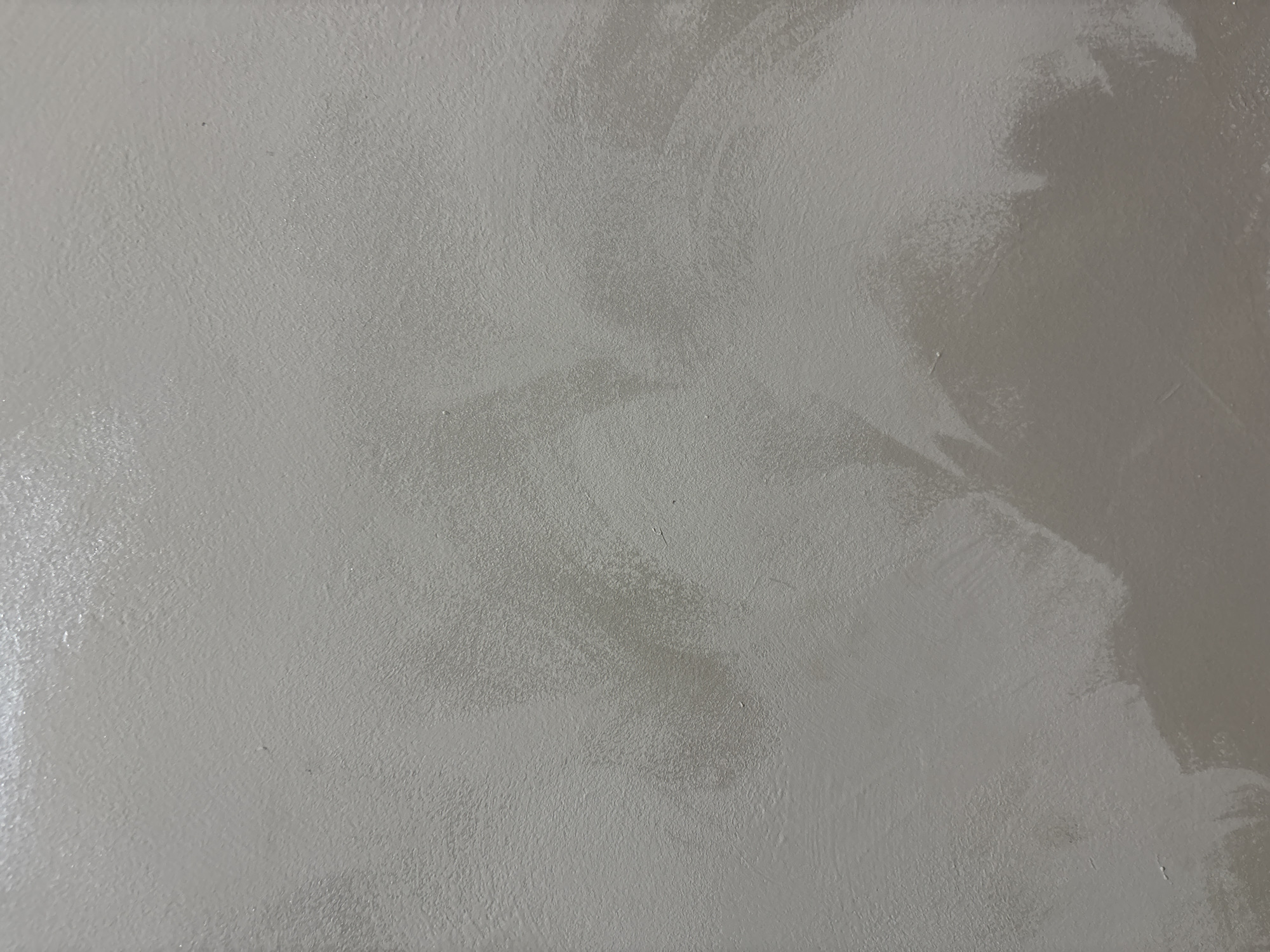 The image shows a close-up, top-down view of a textured, light gray surface. The texture appears to be that of painted plaster or concrete, with subtle variations in sheen and small imperfections visible. There are areas where the paint application is uneven, creating a slightly mottled or streaky effect. Some parts of the surface are lighter and more reflective, suggesting a glossier finish or a different angle of light, while other areas are a matte, more muted gray. The lighting creates soft shadows and highlights, emphasizing the uneven texture. No people, specific objects, or identifiable features of Burgas, Bulgaria are present in the image. The scene is a generic close-up of a wall or ceiling texture.