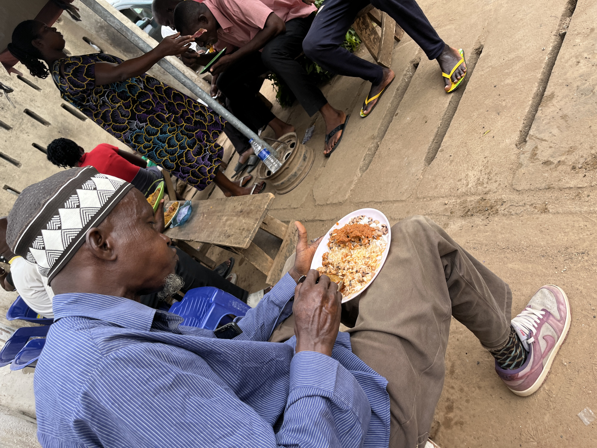 The image captures a casual outdoor gathering in Abuja, Nigeria, centered around people eating and socializing. In the foreground, an older man with dark skin is seen in profile, wearing a black and white patterned cap, a blue striped long-sleeve shirt, and brown pants. He holds a white oval plate with both hands, eating a meal of rice, black-eyed beans, and a reddish sauce with a spoon. His feet are visible, wearing purple Nike sneakers with patterned socks.
In the mid-ground, a woman stands on the left in a long, dark dress with vibrant purple, yellow, and blue floral patterns. Partially obscured behind her is a person in a red shirt. To the right, several men are visible; two are seated or squatting, engaged with mobile phones. One of these men wears black flip-flops with "GUCCI" written on them.
The setting appears to be an informal outdoor eating area, possibly a street-side stall or communal space. The ground is a light-colored concrete or paved surface with linear indentations. A metal pole extends vertically, and a car wheel rim is used as a makeshift table, holding a plastic water bottle. Blue plastic stools are scattered around. The lighting suggests it is daytime, possibly overcast or late afternoon. The individuals appear calm and focused on their meals and interactions.