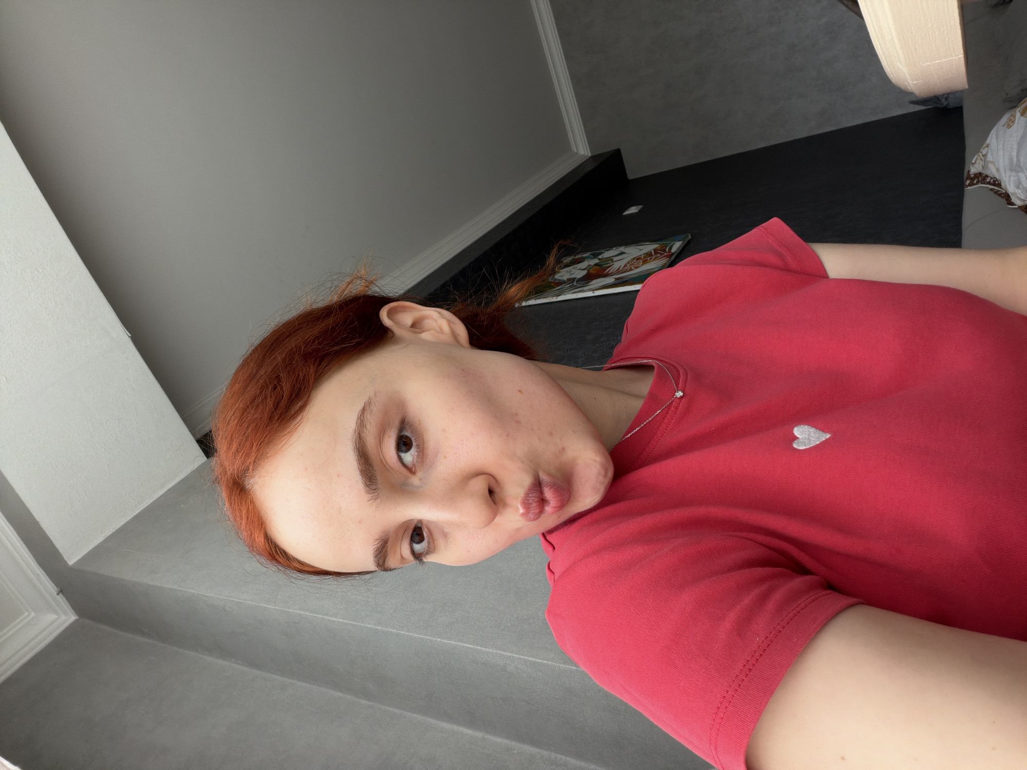 A young woman with red hair and fair skin is posing for a selfie. She's wearing a red t-shirt with a small white heart on the chest. Her lips are pursed into a kiss. The background consists of a gray wall, a black surface with a decorative mat on it, and part of a white object. The location is not discernible from the image alone to confirm the exact place in Tbilisi, Georgia.