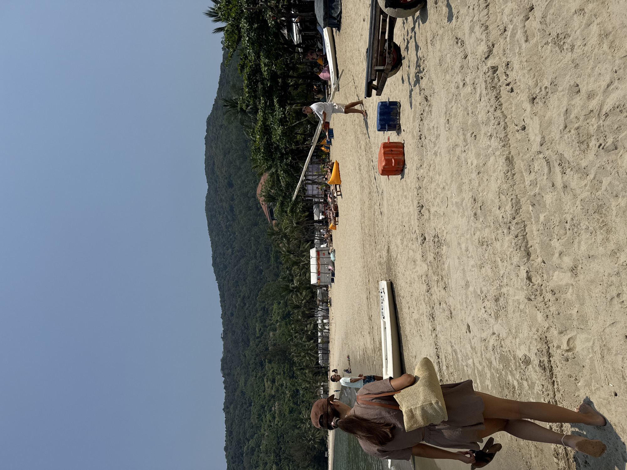 The image displays a daytime scene on a sandy beach, identified as a bay likely on the Son Tra Peninsula, Đà Nẵng, Vietnam. The environment consists of a clear blue sky, light tan sand, calm water, and densely forested mountains in the background.
In the immediate foreground, a female subject, viewed from behind, walks barefoot on the sand, carrying dark sandals in her left hand and a light brown woven bag on her right shoulder. She wears a brown two-piece top and shorts, and a brown baseball cap.
Mid-ground details include a white small boat resting on the sand near the water's edge. A male subject in a light blue t-shirt stands near the water. Further to the right, another male subject in a white and blue striped shirt and white shorts holds a long, light-colored, pole-like object horizontally across his body. An orange and a blue buoyant cube are positioned on the sand near him, alongside a black two-wheeled trailer on the far right.
The background shows calm, dark blue-green bay water. The beach is lined with numerous palm trees, behind which several white tent-like structures are visible. Multiple individuals are scattered across the beach, some relaxing on yellow inflatable loungers or sunbeds.