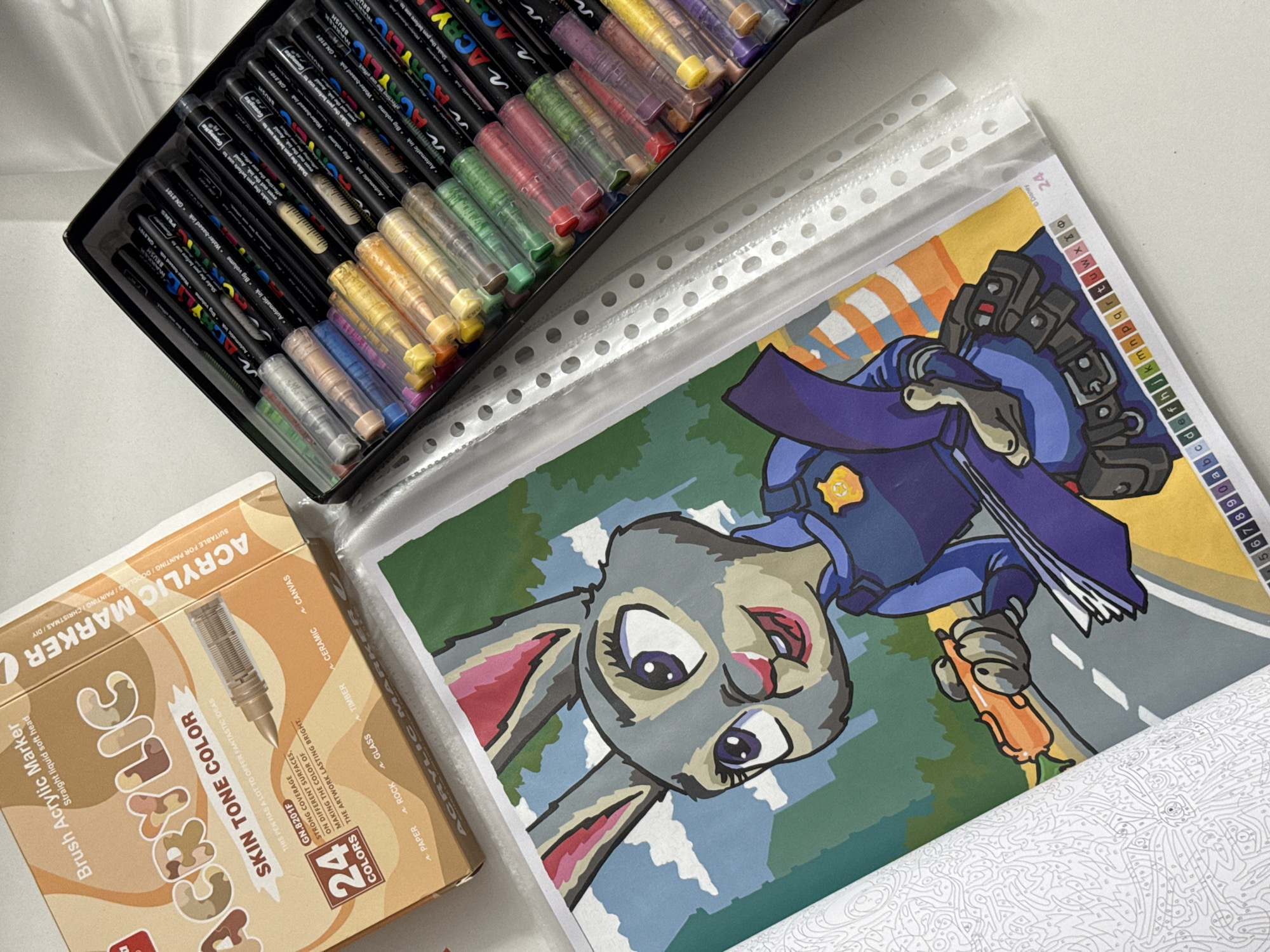 The image displays a flat lay of art supplies, including a box of "Brush Acrylic Marker Straight liquid soft head" with a focus on "SKIN TONE COLOR" and "24 COLORS", and a case of assorted acrylic markers in various colors. A partially completed paint-by-number page featuring a cartoon rabbit character in a police uniform holding a carrot is visible in the foreground. The scene is set on a white surface, suggestive of a table or desk. There is no information or visual context to suggest any specific location in the city of Łódź, Poland. The image focuses solely on the art materials and the painting in progress.