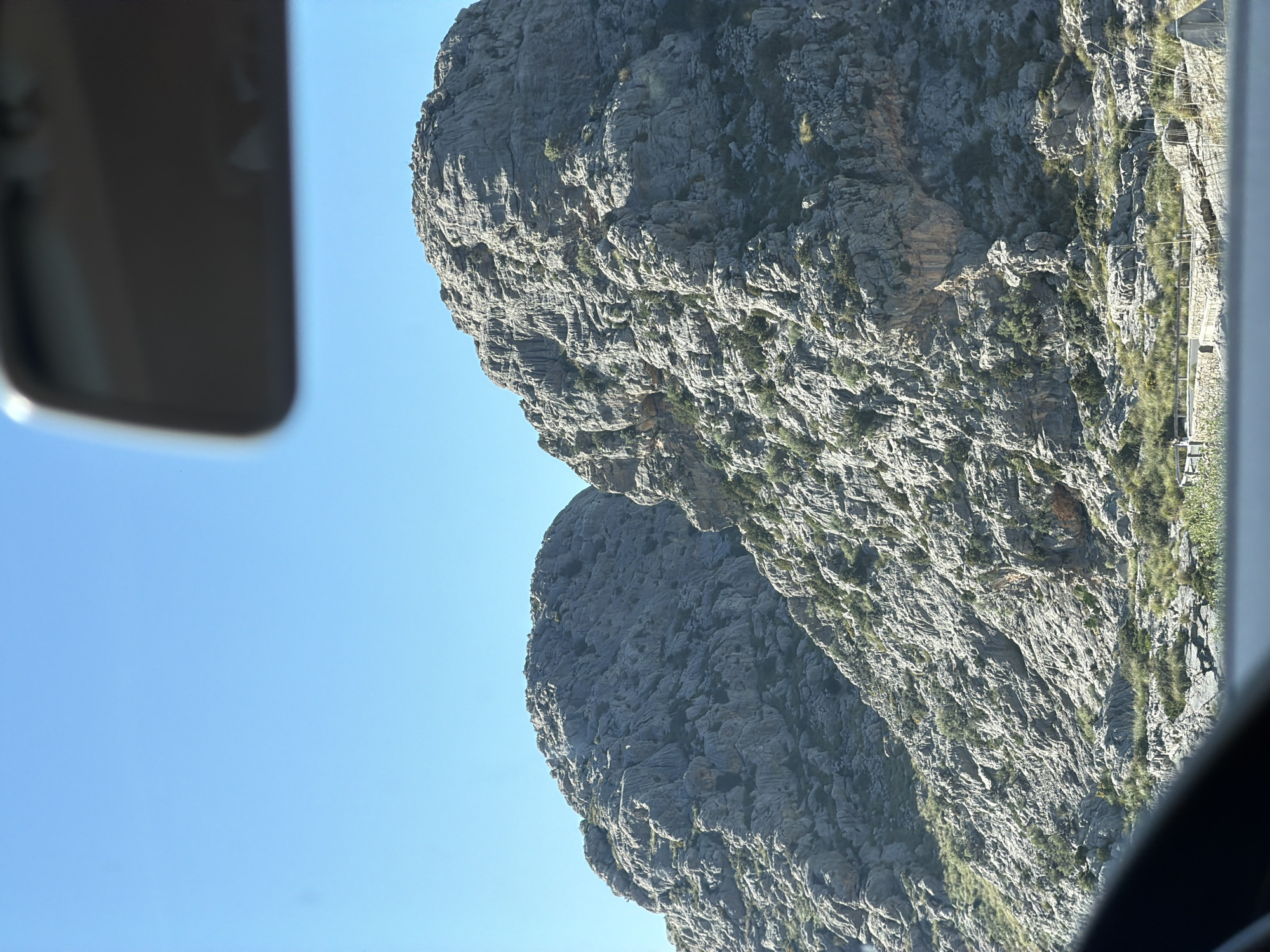 The image shows a rocky mountain landscape taken from inside a vehicle. Part of the vehicle's interior, including a sun visor, is visible on the left side of the frame. The mountain appears steep and rugged with visible rock formations and some vegetation. The sky is clear and blue in the background. The location is the municipality of Escorca, Spain, based on the landscape characteristics. The image captures a moment of travel through the mountainous terrain.