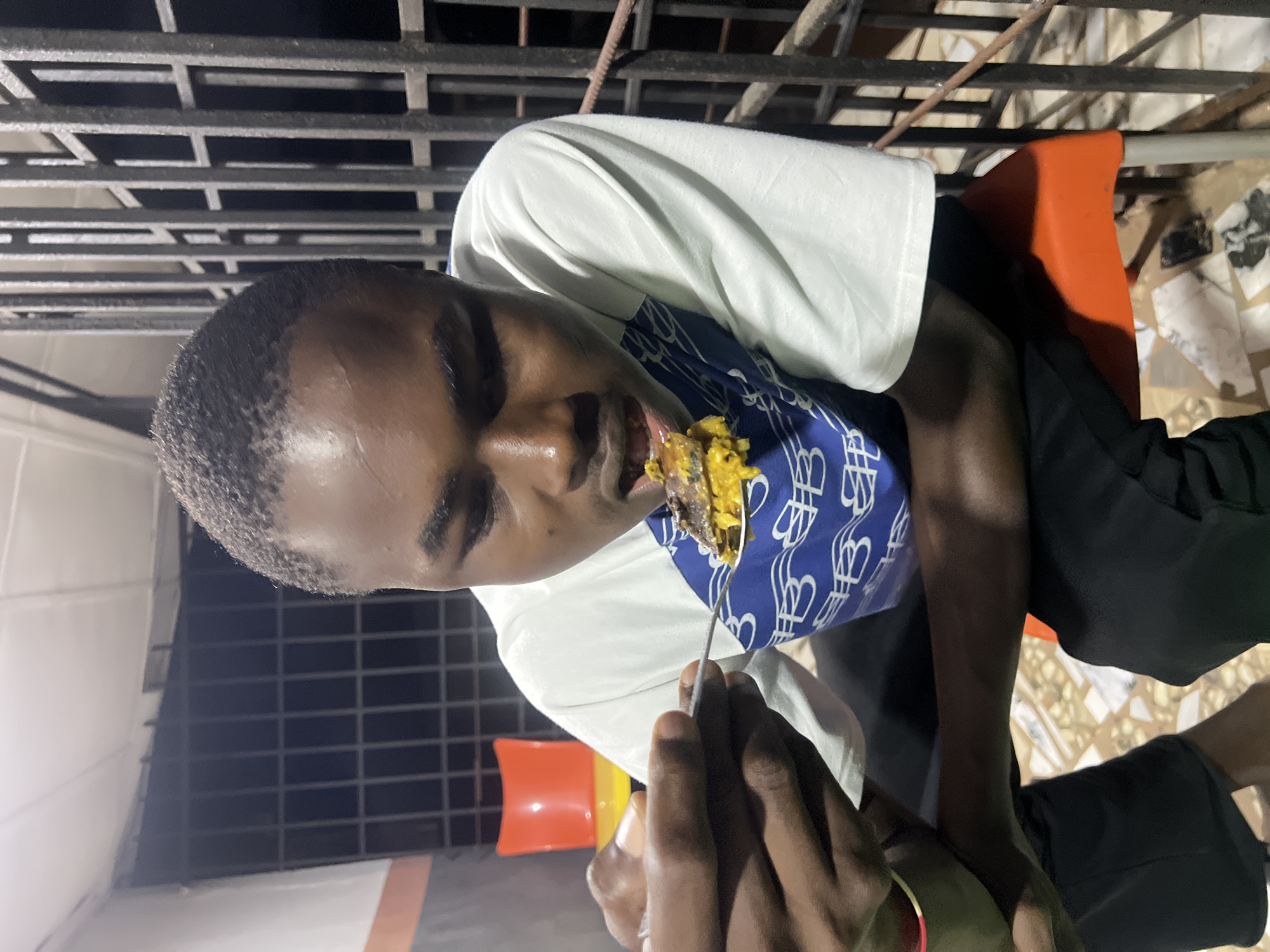 A young Black male is captured in a medium shot, indoors, in Nkpor, Nigeria, as he brings a spoonful of food to his mouth. He is seated on an orange plastic chair with metal legs. He is wearing a white long-sleeved shirt with a blue graphic print on the chest. His head is tilted down, focusing on the food. His left hand is visible, holding a silver spoon with a portion of yellow food, possibly rice and stew, and a piece of meat. The background features a black metal grate or fence, suggesting an outdoor or semi-outdoor setting. To the left, partially visible, is a red plastic chair with yellow accents. The lighting appears to be artificial, casting shadows and highlighting the subject's face and hand. The floor is tiled with a mosaic pattern.