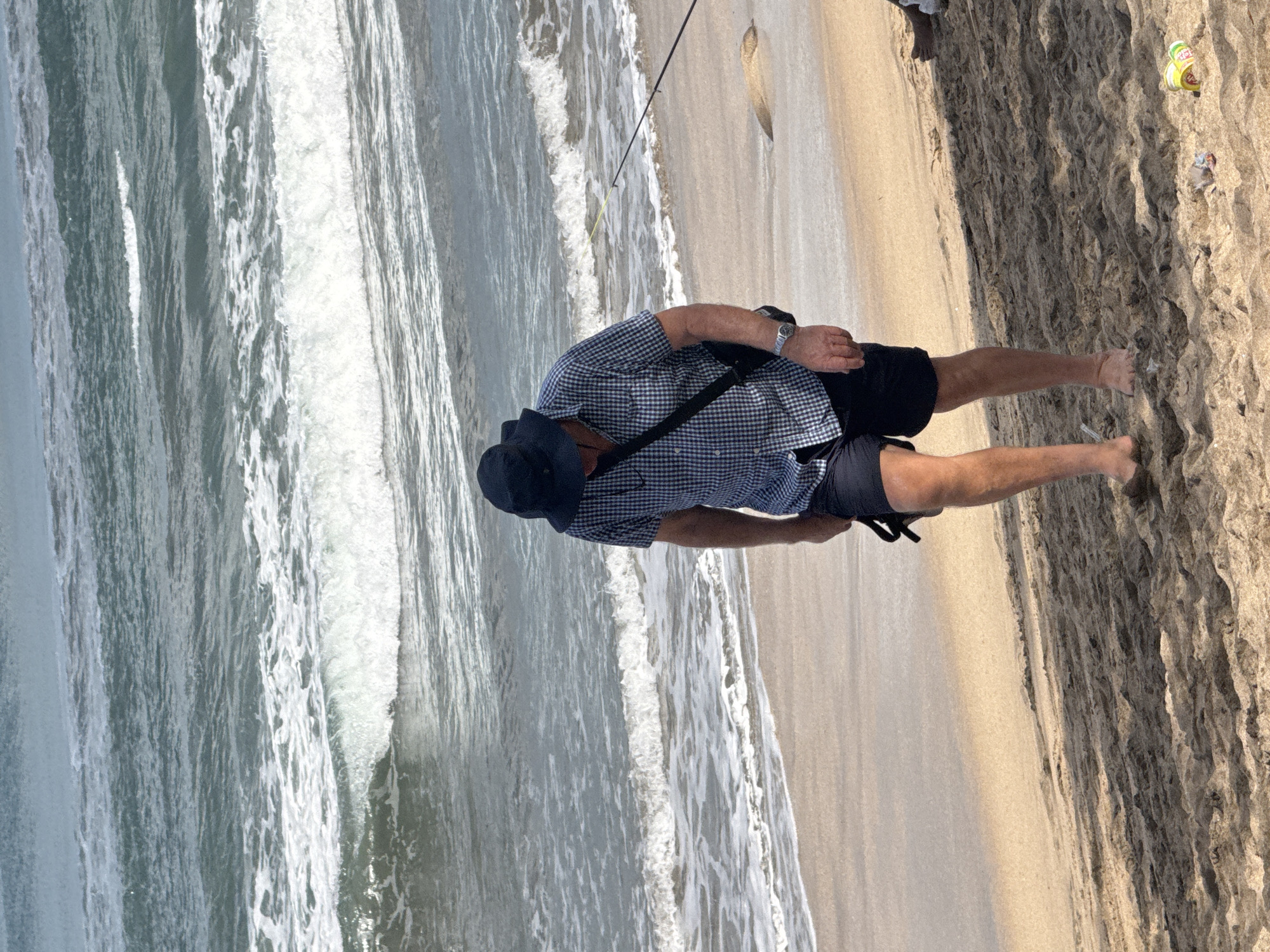 A man wearing a blue and white checkered short-sleeved shirt, black shorts, a black hat, and a black crossbody bag walks down a sandy beach in Kuta, Indonesia, towards the ocean. The waves are breaking on the shore. A fishing rod is visible in the background. There is some litter on the sand.