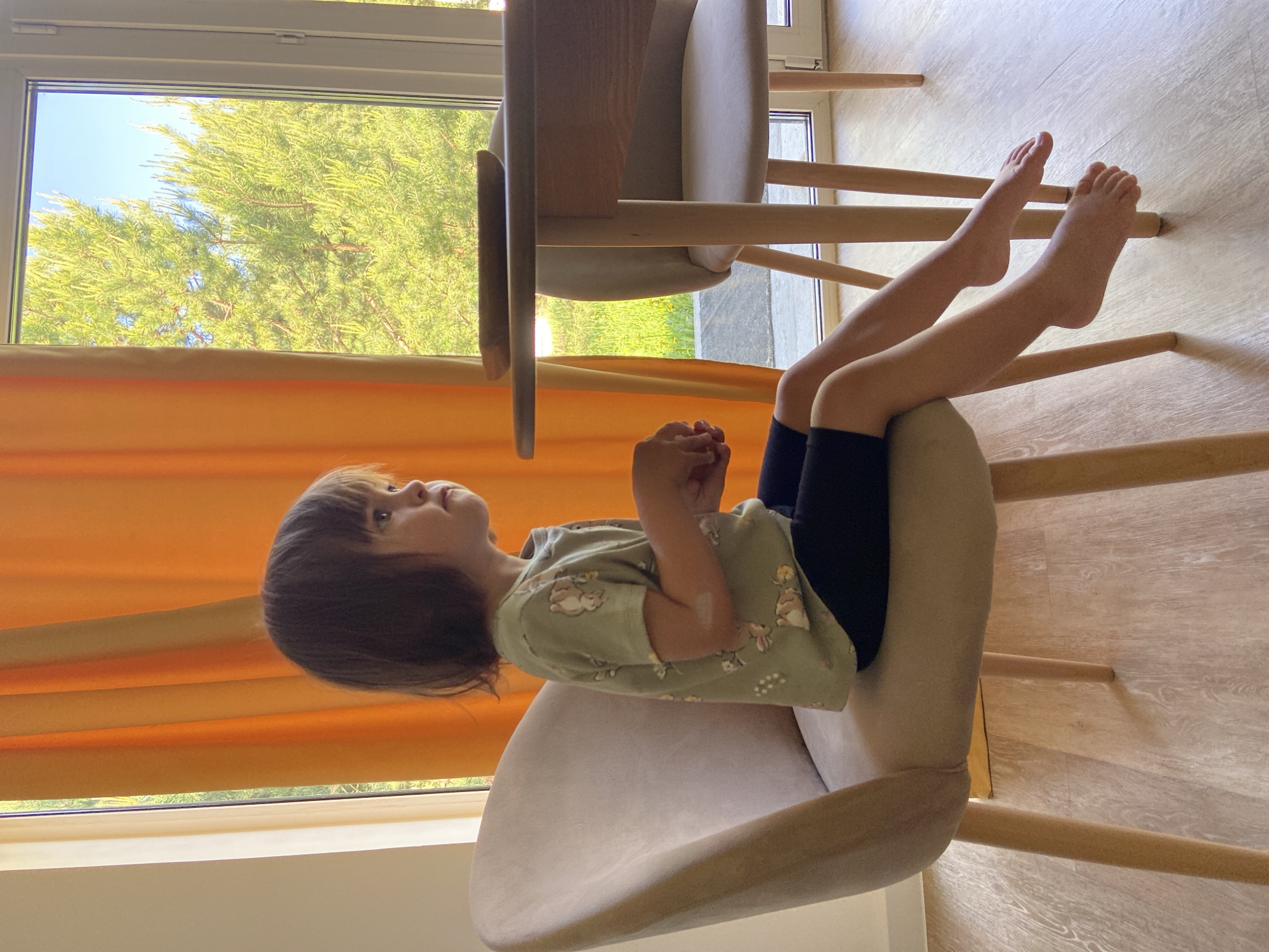 A child is sitting backwards in a chair. They are wearing a green shirt with animal patterns and black shorts. Their legs are extended forward and resting on the back of another chair. The chairs are light-colored wood with padded seats. A window is visible in the background with orange curtains partially drawn. Trees are visible outside the window. The floor is a light wood-patterned material. The scene appears to be indoors, likely in a home. It is not possible to determine the location as Tyumen, Russia, from the image alone.