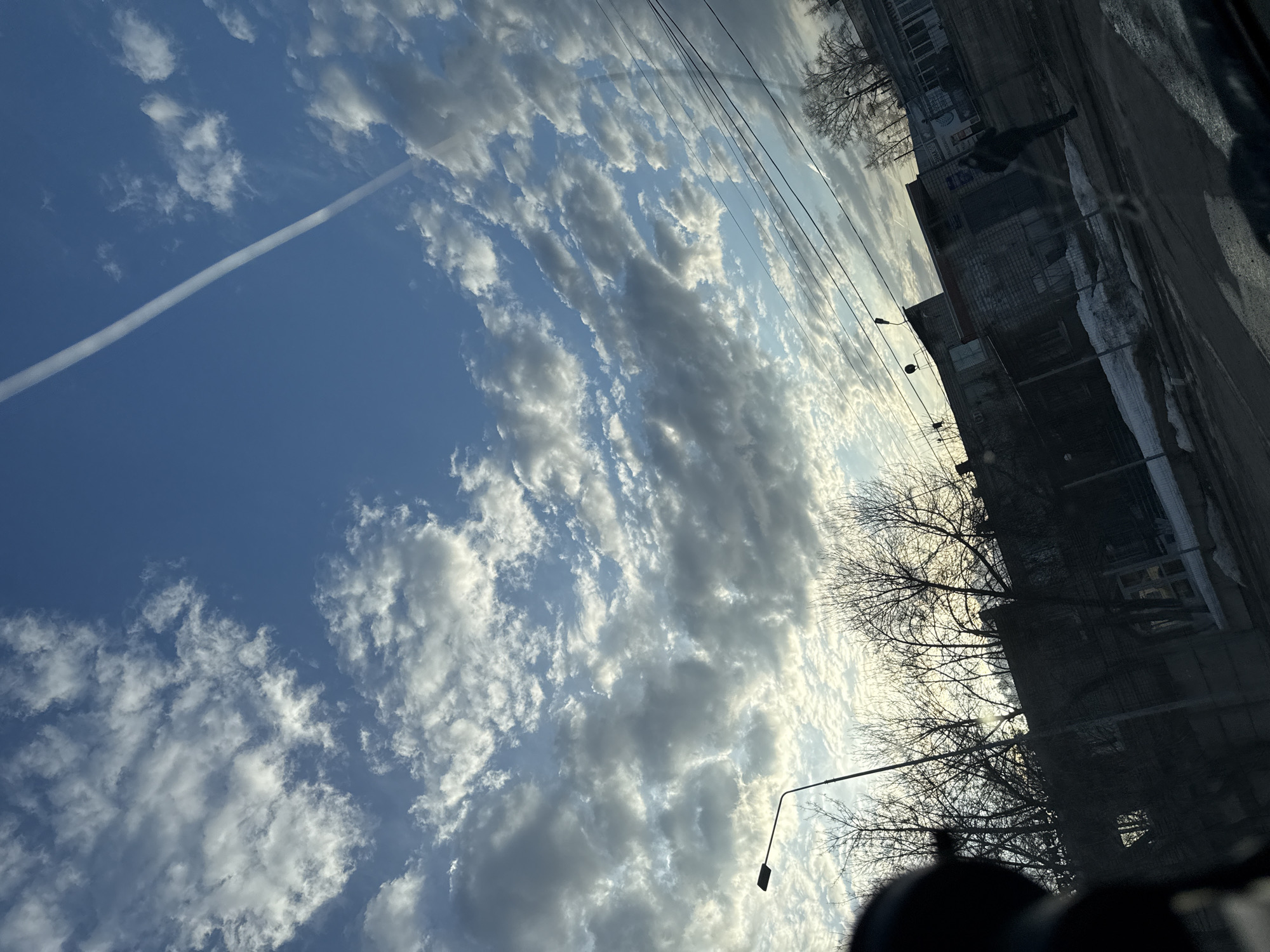 The image shows a view from inside a vehicle, possibly a car, in what appears to be a city environment. The sky occupies a significant portion of the image, displaying a vibrant blue color interspersed with numerous white, fluffy clouds. A distinct contrail stretches across the blue expanse, indicating the passage of an aircraft.
On the right side of the image, a street scene is visible, albeit partially obscured by reflections on the car's window. Buildings with a weathered, possibly Soviet-era architectural style line the street. Bare trees, typical of late autumn or early winter, are interspersed between the buildings. A street light is visible. Patches of snow and puddles can be seen on the road, further suggesting a cold season.
The reflections on the window make it difficult to discern exact details, but it appears a person is walking, but it is hard to tell which way.
Without precise location data, pinpointing the exact street in Chaikovsky, Russia, is impossible. However, the architectural style and overall atmosphere suggest a typical residential or commercial area within the city.