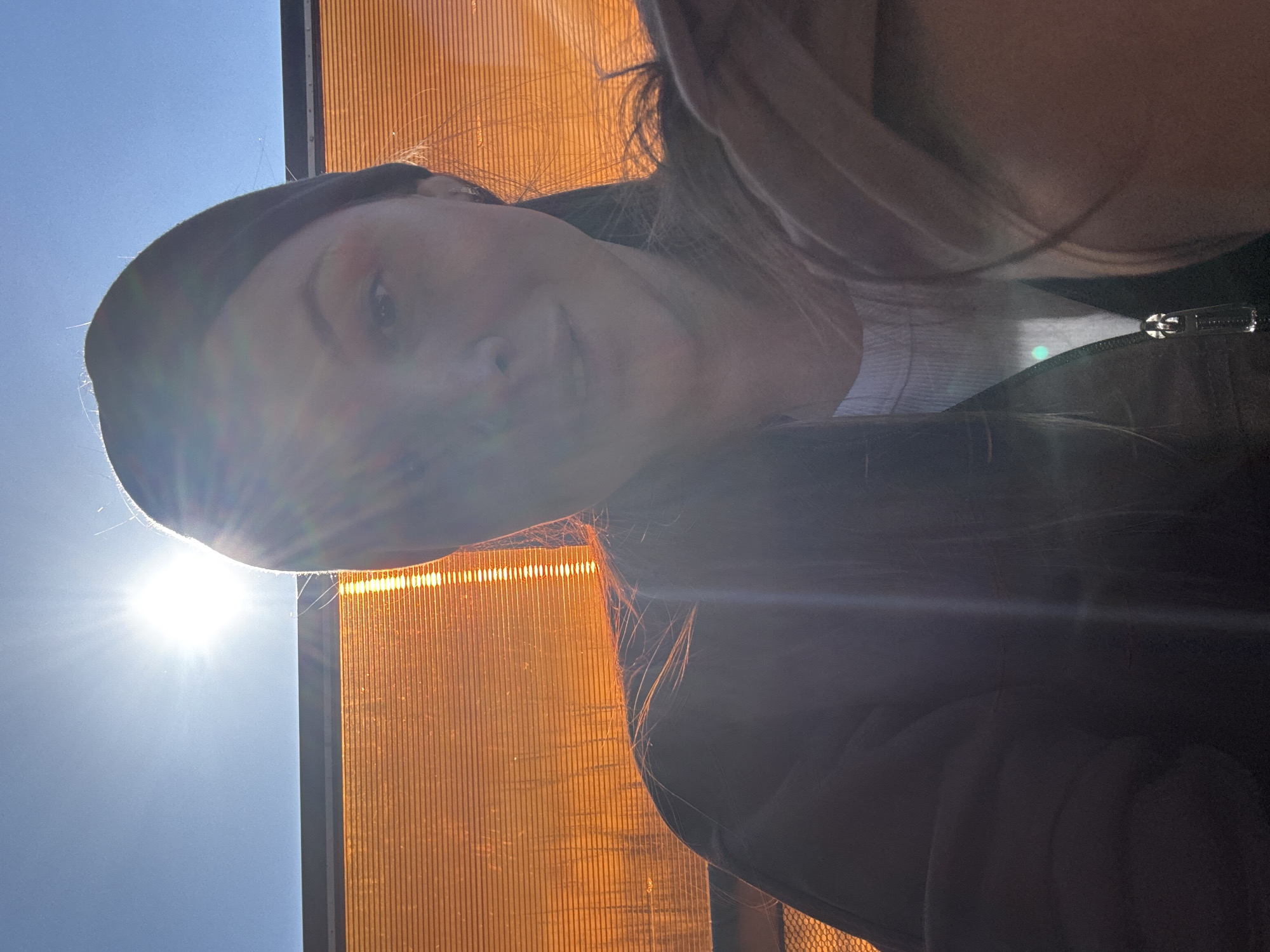 A woman is taking a selfie in bright sunlight. She is wearing a black headband and a black jacket. The background consists of a blue sky and an orange vertical-striped structure. The location is Кипчак, Russia.