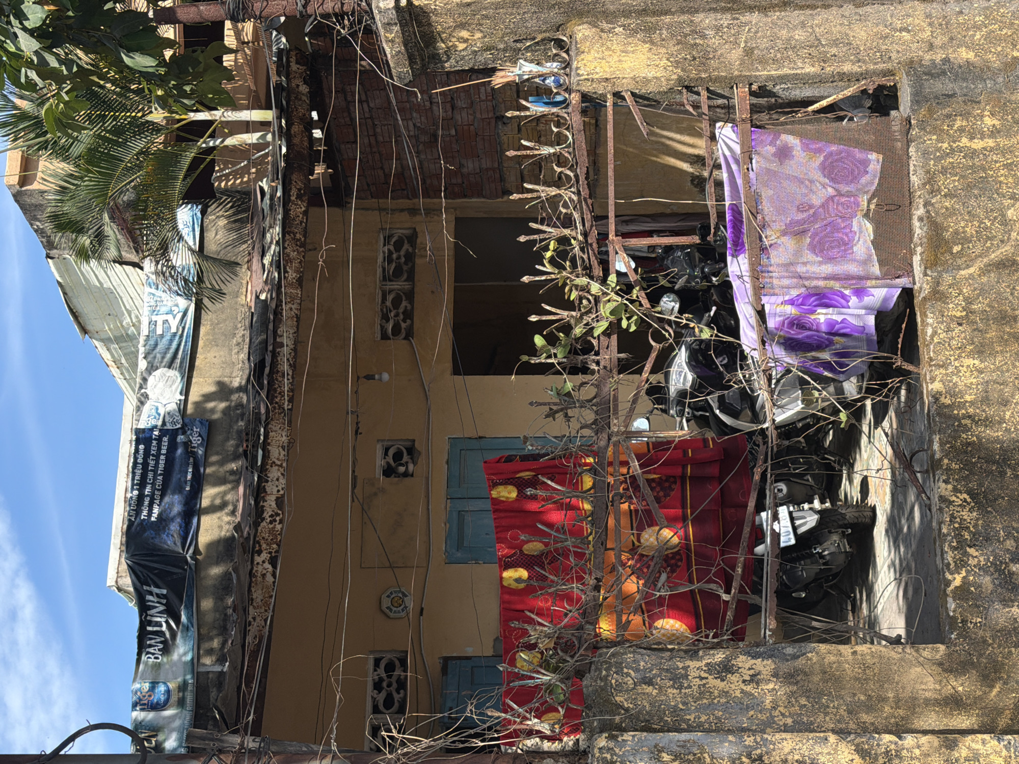 This image shows a decaying building exterior in what is likely Đà Nẵng, Vietnam. The building has faded beige walls with old, patterned windows framed by blue shutters. A rusted metal gate covers a portion of the building's facade. Inside the gate are two motorcycles, partially obscured by fabrics and possibly a blanket with floral patterns and a red fabric with orange fruit. The exterior walls are marked with weathered signage. Power lines and overgrown branches add to the building's state of disrepair.