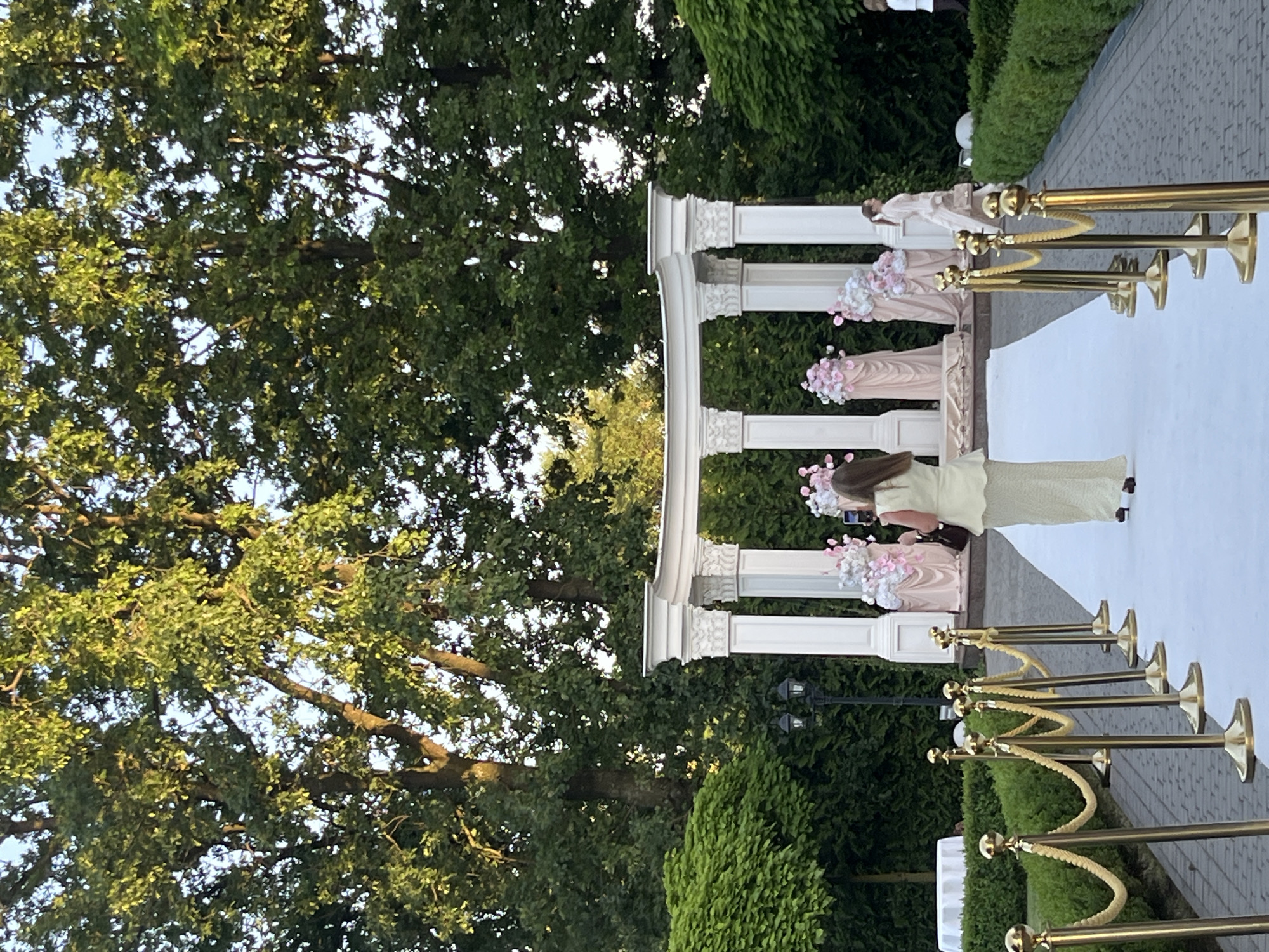 The image depicts an outdoor wedding ceremony setup in the city of Dubivtsi, Ukraine. A white carpeted aisle is laid out, flanked by gold stanchions and ropes, leading to a white neoclassical gazebo adorned with floral arrangements. A woman in a light-colored dress is standing on the carpet, facing the gazebo and holding a mobile phone, appearing to take a photograph or video. In the background, several other individuals are visible, including one in a light-colored dress walking away from the gazebo, and another person further in the distance. The setting is characterized by lush green trees and manicured hedges, suggesting a well-maintained outdoor venue. The overall scene suggests a formal event or celebration taking place in a park-like environment.