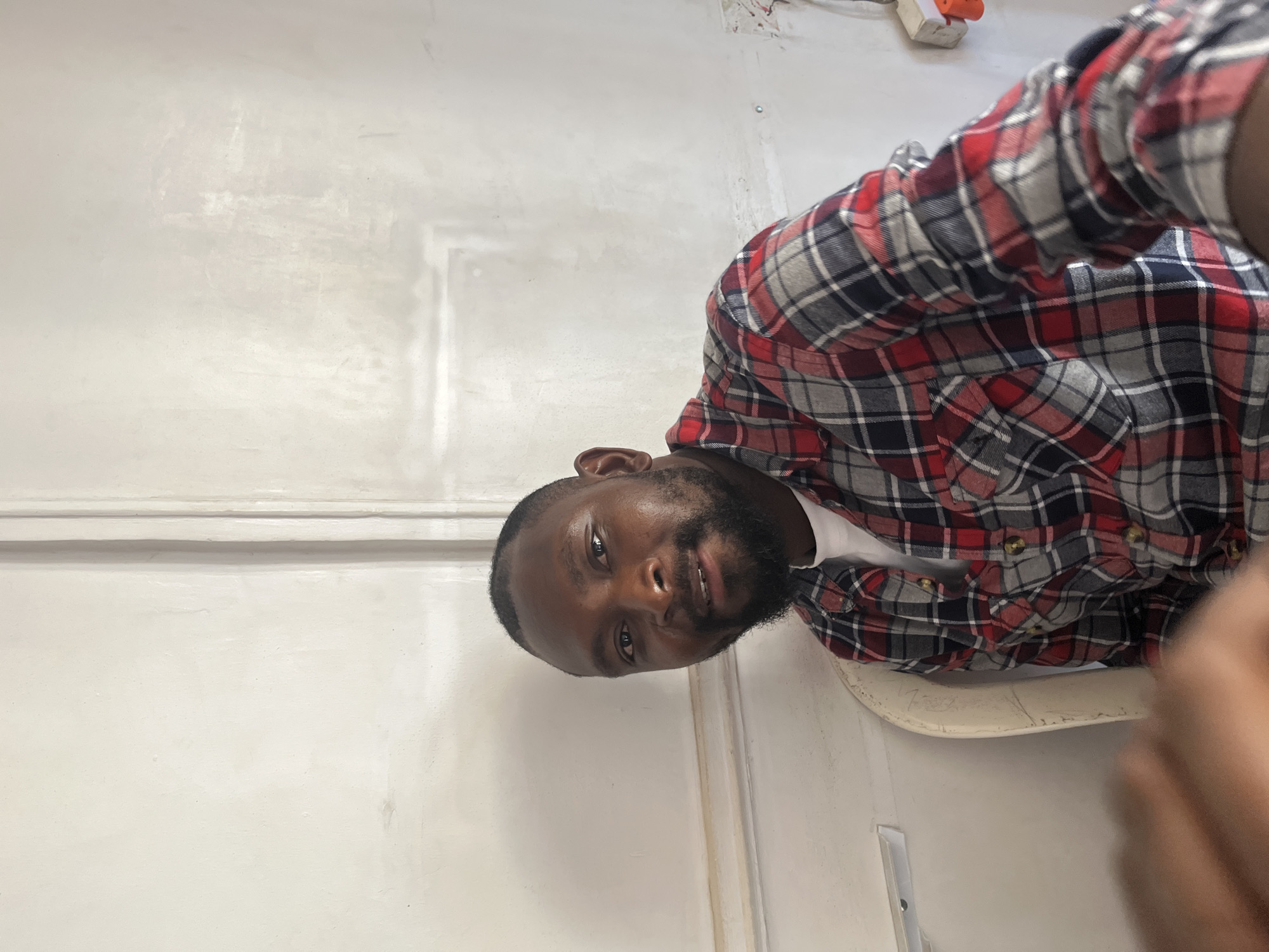 A person with dark skin and a beard is visible, wearing a red, black, and gray plaid shirt with brown buttons. They are positioned diagonally against a plain, off-white wall, likely indoors. The wall has a textured molding near the top and a vertical seam or line running down it. A portion of a white chair is visible. The person is likely in a residential building in Jos, Nigeria.