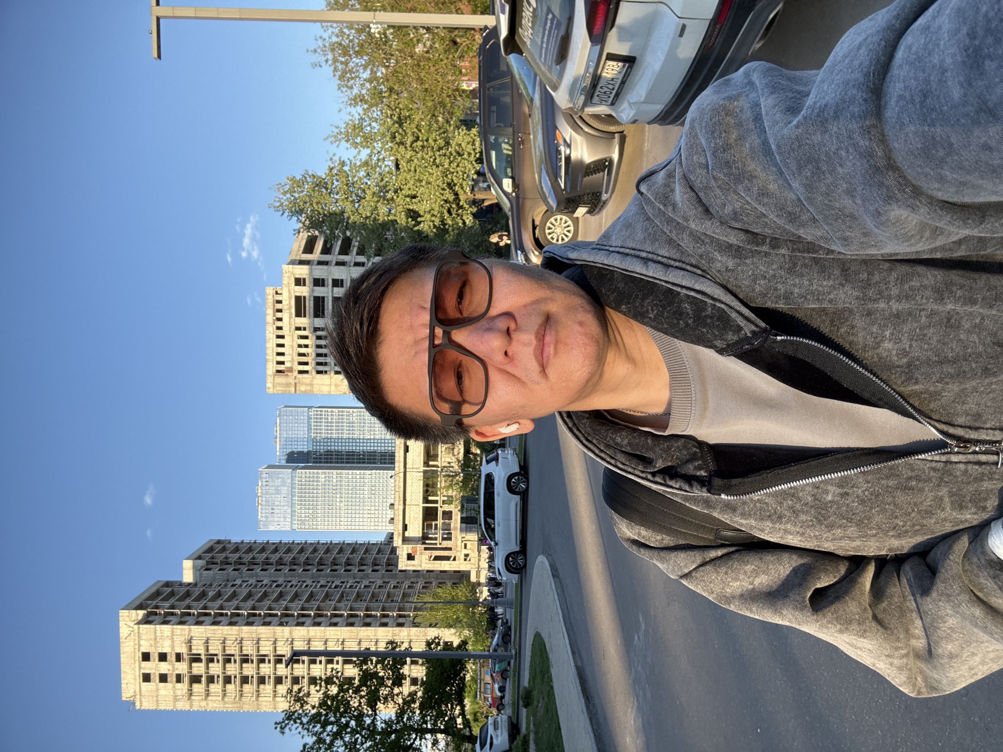 A middle-aged man wearing glasses, a gray jacket, and an earpiece is taking a selfie. In the background are buildings under construction and completed high-rise buildings, indicating an urban environment. Cars are parked along the side of the road, and the setting suggests an ordinary street scene in a city. Based on the license plate visible on one of the cars, the location appears to be Moscow, Russia.