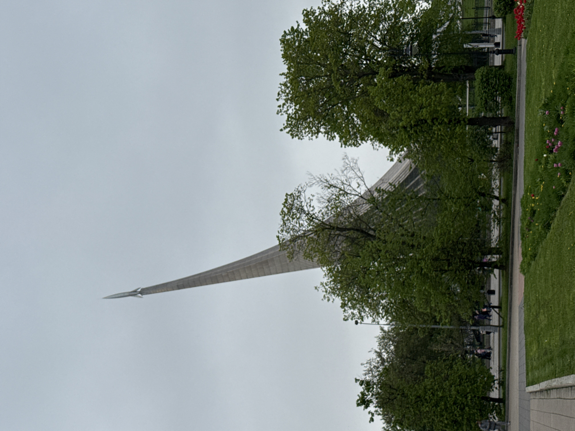 The image shows the Monument to the Conquerors of Space in Moscow, Russia, partially obscured by trees. The monument, a large obelisk topped with a model of a rocket, is set against a grey, overcast sky. The base of the monument is visible through the trees, and part of a park with flowers and a walking path is in the foreground. People are sitting on benches along the path.