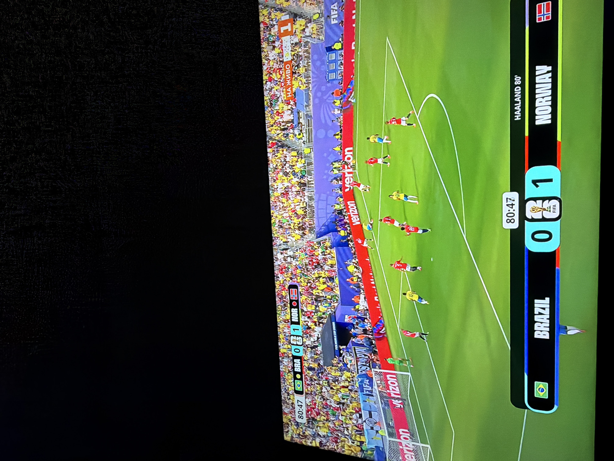 The image shows a television screen displaying a live soccer match between Brazil and Norway. The score is 0-1 in favor of Norway. The match is in its 80th minute, and a goal was scored by Haaland. The stadium is packed with spectators wearing yellow and green, the colors of Brazil. The environment is a soccer field with white lines marking the boundaries. The image does not provide any information about the city of Ognianovo, Bulgaria.