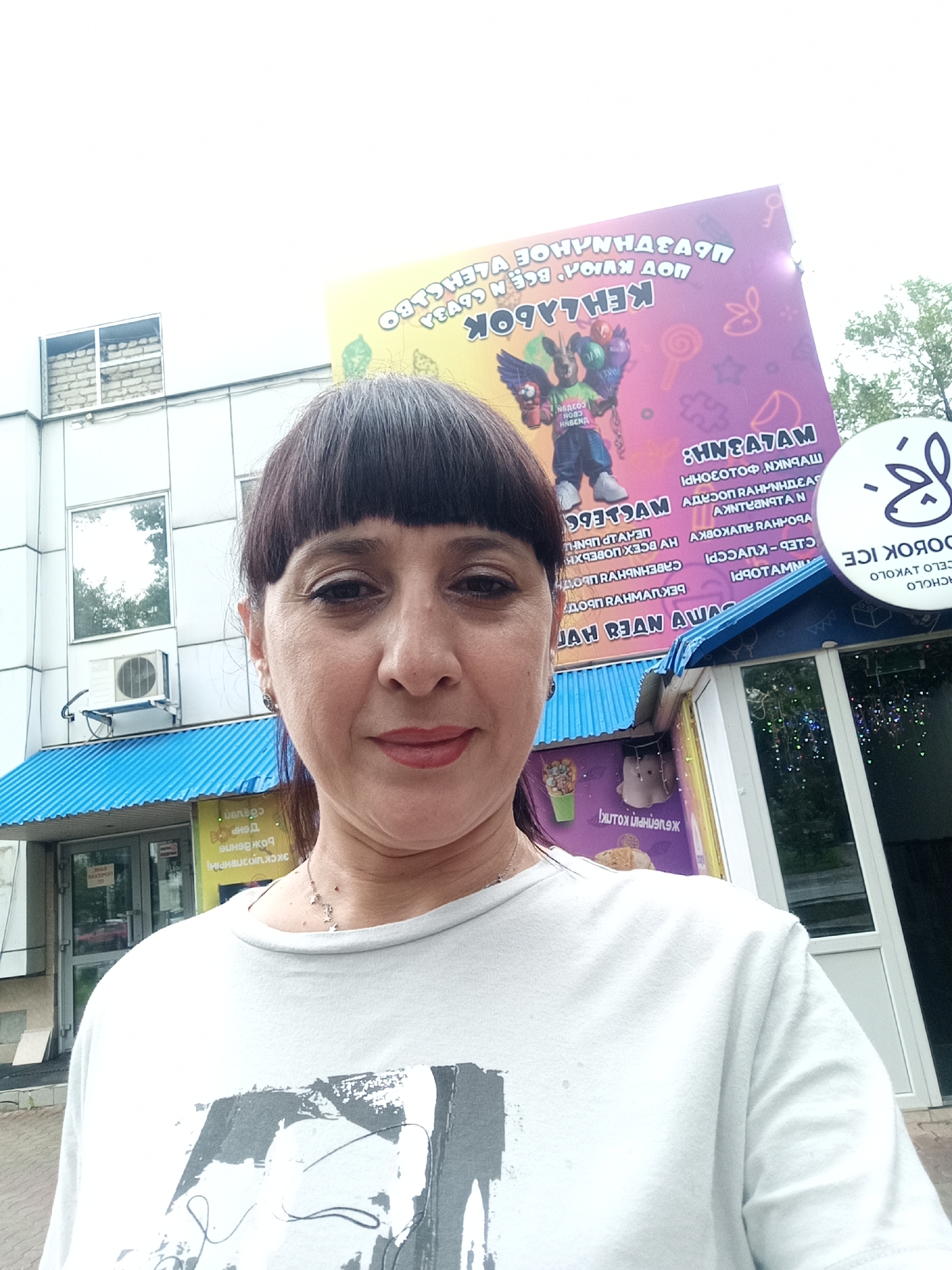 A woman with dark hair and bangs is standing in front of a building with a blue awning. She is wearing a white t-shirt with a graphic print on it. Behind her, a large, colorful banner displays text in Russian, advertising "Ice Cream" and "Kids Fun" and features an illustration of a winged creature. To the right of the banner, a circular sign with a rabbit logo reads "Dorogoe Ice". The building appears to be a commercial establishment. The background shows an urban setting with trees and a cloudy sky.