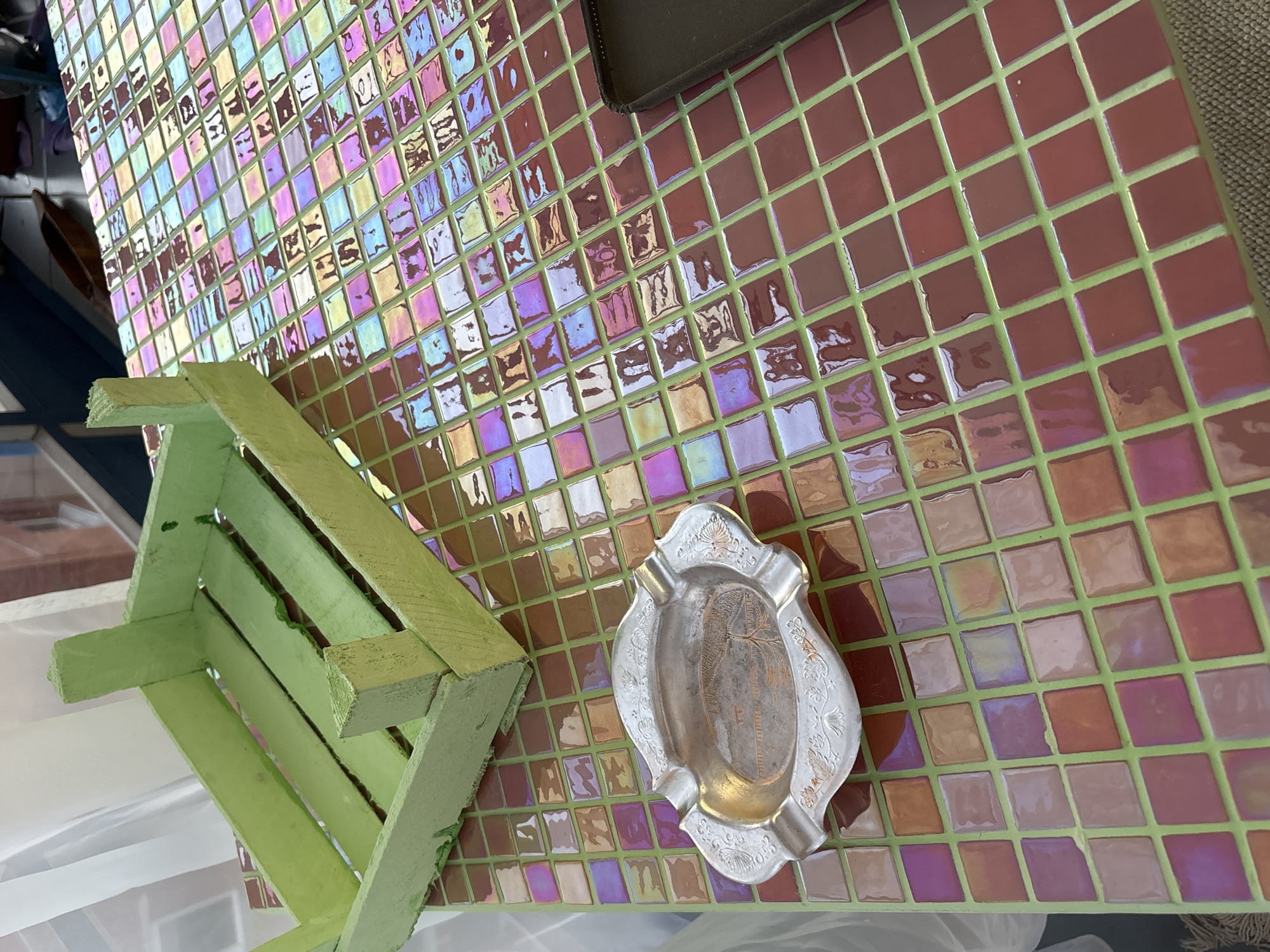 The image provides a close-up, high-angle view of a surface covered with iridescent mosaic tiles. The small, square tiles are predominantly reddish-brown or maroon, but their glossy finish reflects a vibrant range of colors including pink, purple, gold, green, and blue. The grout lines separating these tiles are a distinct light green.
In the lower right section of the frame, a decorative metal ashtray is prominently displayed. It is silver-colored with intricate, raised patterns along its scalloped edges and a central oval area featuring a gold-toned design, possibly depicting a stylized palm tree or similar motif. The ashtray has several indentations around its rim for resting cigarettes.
To the left, a light green wooden crate is partially visible, resting on or adjacent to the tiled surface. The crate appears rustic, with visible slats and a pastel green paint finish that shows some signs of wear. In the upper right corner, a dark, rectangular object, possibly a wallet or phone case, is partially visible, also situated on the tiled surface.
The background to the far left is out of focus, revealing vague shapes that suggest a building or window, with hints of blue and reddish-brown structures and some white material below the crate. The scene is brightly lit, likely by daylight, which accentuates the reflective properties of the tiles. No specific activity or event is taking place, and no text is visible within the image.