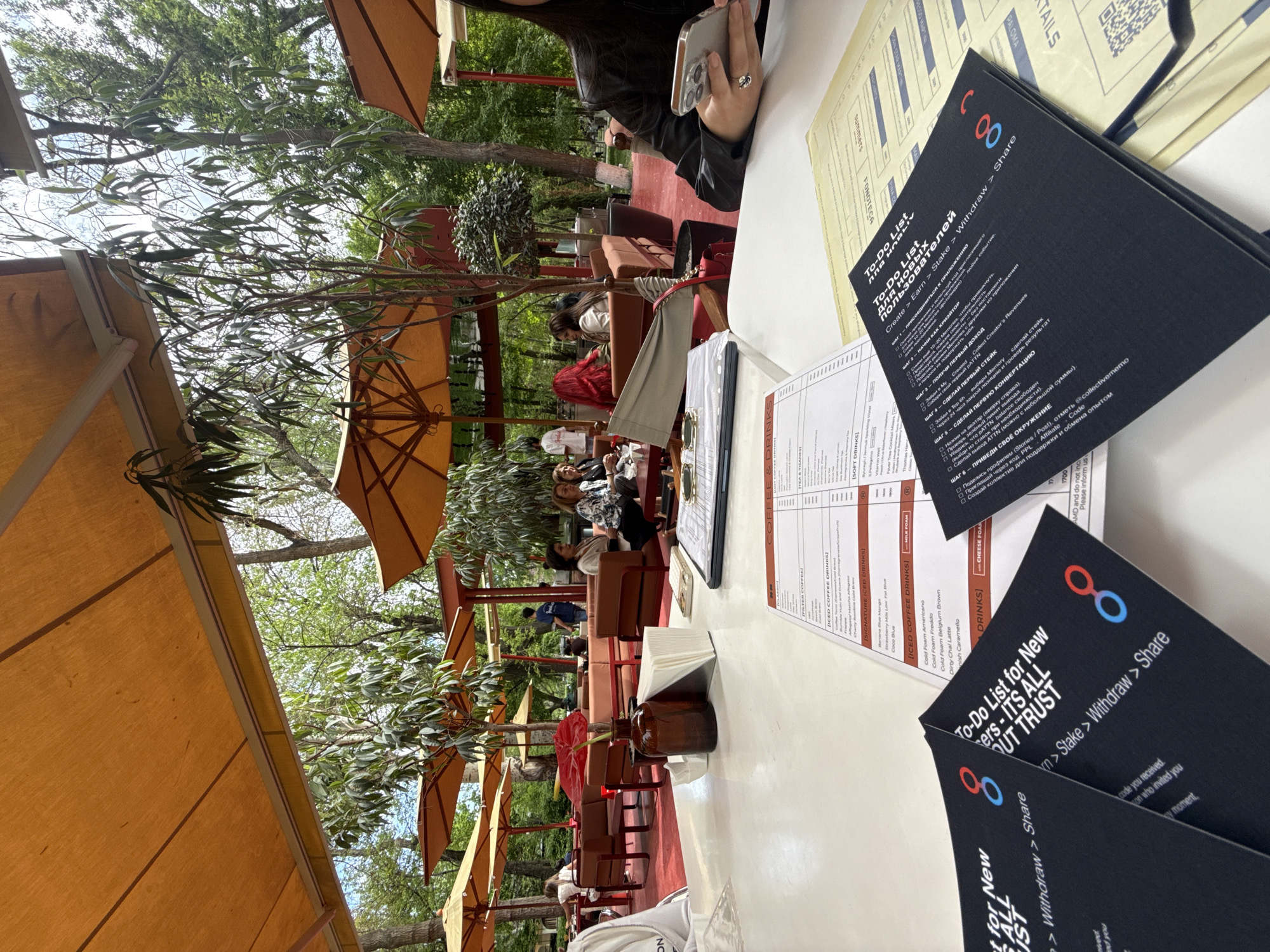 The image depicts an outdoor cafe in Yerevan, Armenia. Patrons sit at tables shaded by orange umbrellas. Menus and "To-Do List" cards are on the table. One individual is holding a cell phone. Plants decorate the outdoor seating area.