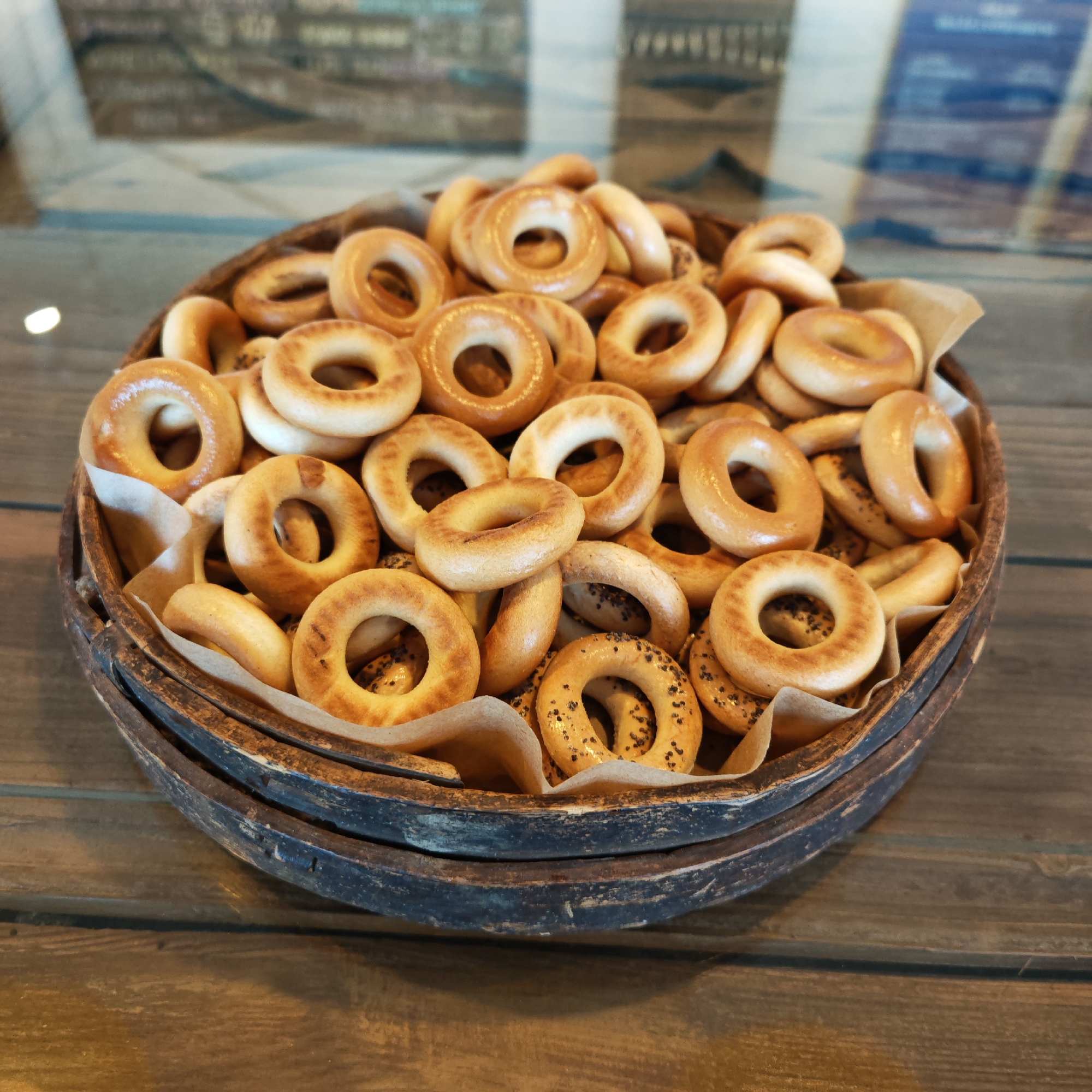 A close-up, medium-shot captures a wooden bowl filled with round, bagel-like pastries. Some pastries are plain, while others have black specks on them. The bowl has two tiers and sits on a wooden surface. The background is blurred, but there's a hint of reflective glass and a slightly visible interior space. This image was taken in Rostov, Russia.