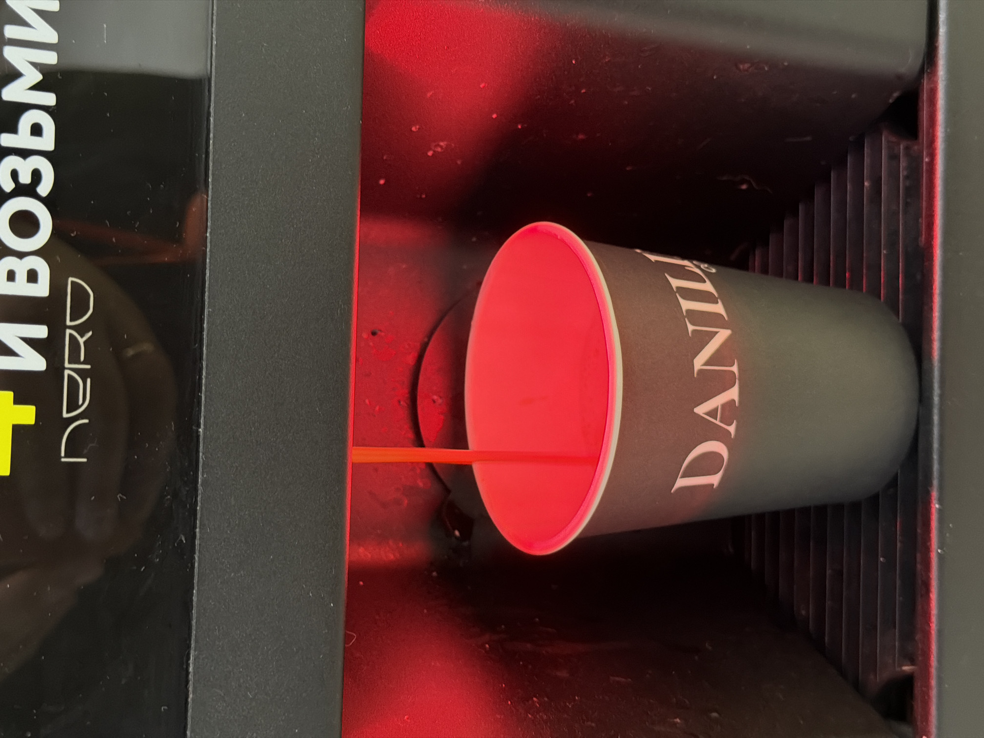 The image shows a close-up, eye-level view of an automatic coffee machine dispensing coffee into a dark-colored paper cup. The coffee stream is a bright orange, illuminated by a red light emanating from within the machine. The cup has the word "DANIIL" printed in white lettering. The coffee machine is black, with some text visible at the top which appears to be in Cyrillic letters: "И ВОЗЬМИ" and "NERO". The coffee machine is situated on a shelf-like structure with horizontal ridges. The overall impression is that of a commercial establishment, possibly a cafe or a shop, in Chaikovsky, Russia, where such machines are common.