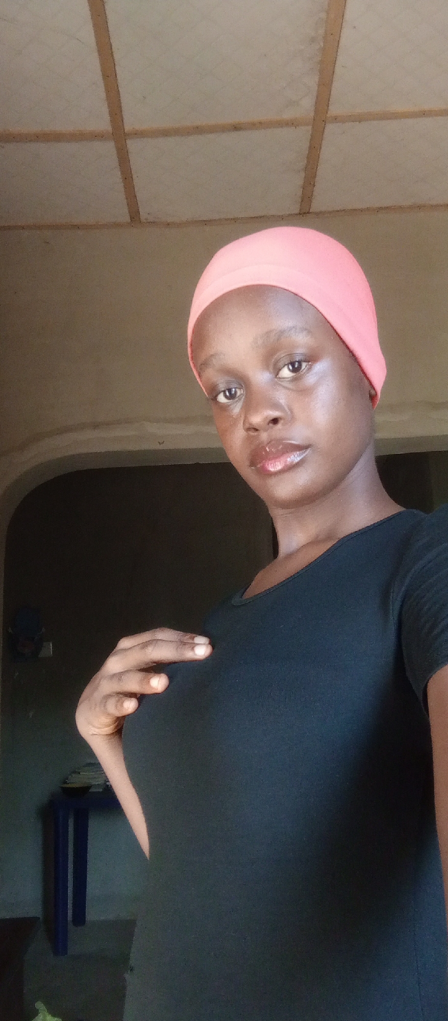 A young woman with dark skin, wearing a peach-colored headscarf and a black shirt, is posed indoors. She is looking towards the camera and has her right hand on her upper chest. The background shows a room with an archway leading to a darker area. To the left, there is a small table with a dark object on top. The ceiling is made of rectangular panels. The location is Akwanga, Nigeria.